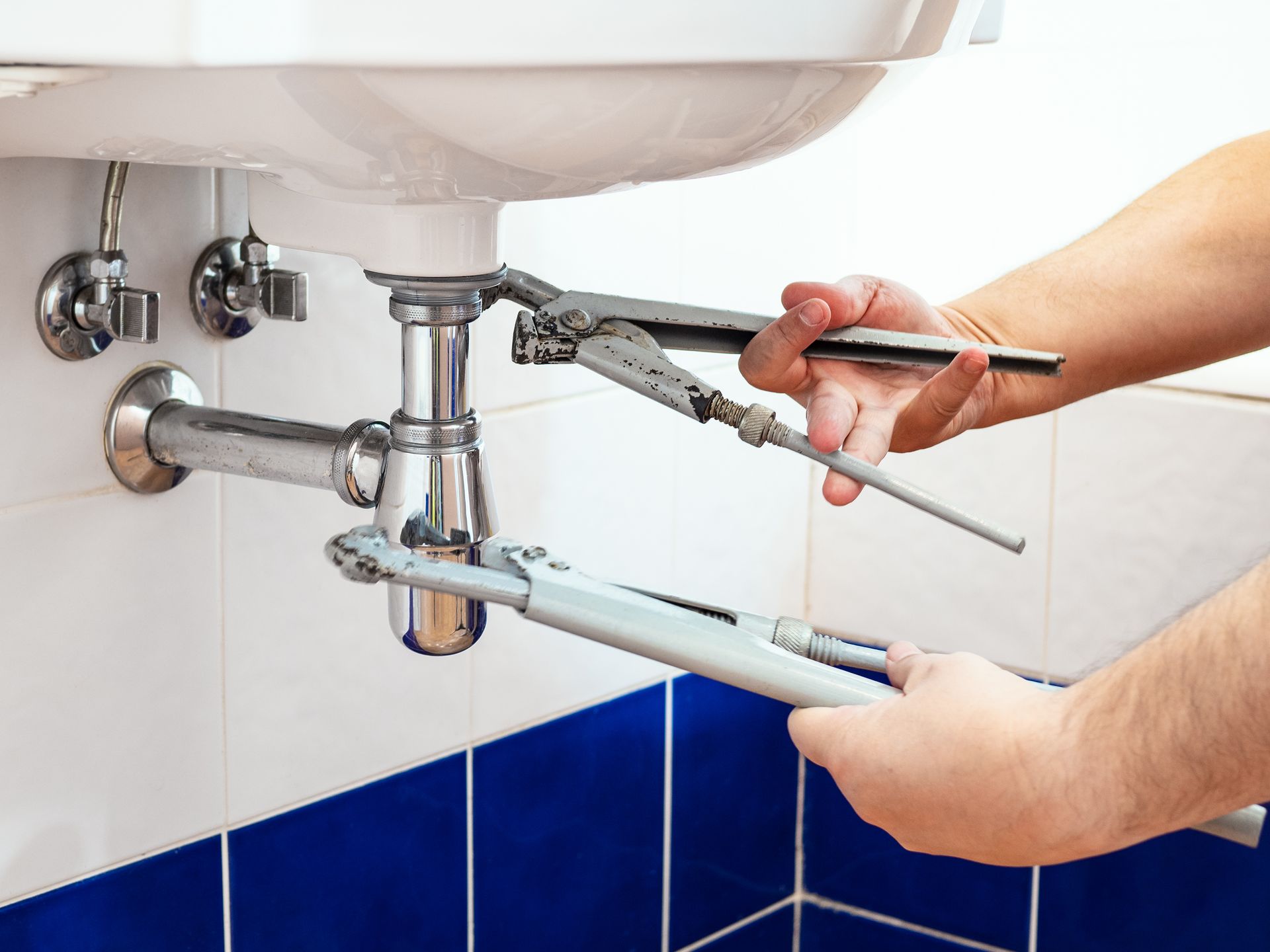 A person using two wrenches to repair a sink drain, with blue and white tiled wall.