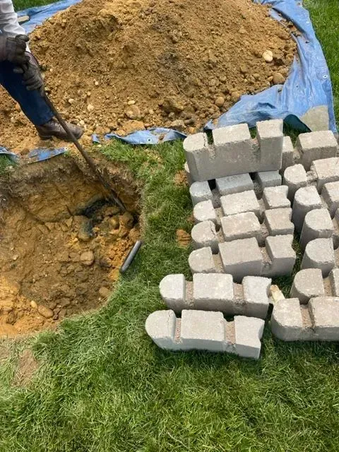 Person digging a hole in the dirt, next to a pile of gray landscaping bricks on grass.