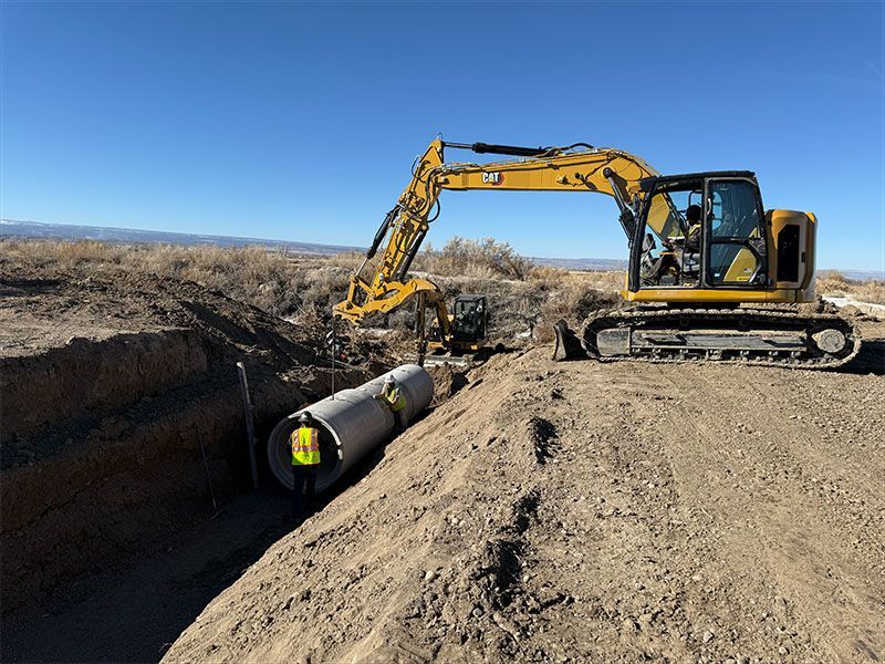 An excavator in a dirt trench is placing a large pipe, on a bright day with a clear sky.