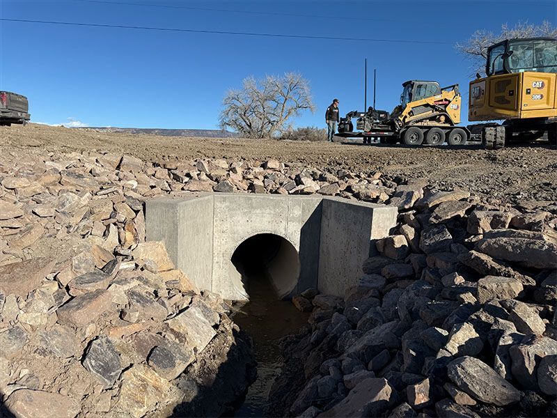 Concrete culvert under a road with riprap, construction vehicles, and workers in a rural setting.