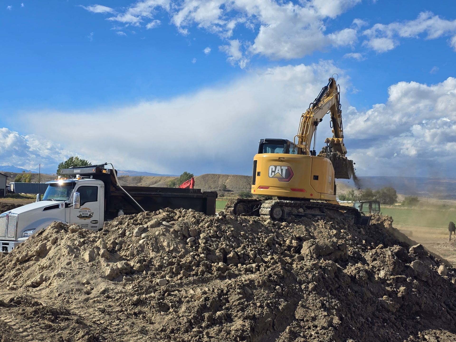 A yellow excavator loads dirt onto a dump truck on a mound under a cloudy blue sky.