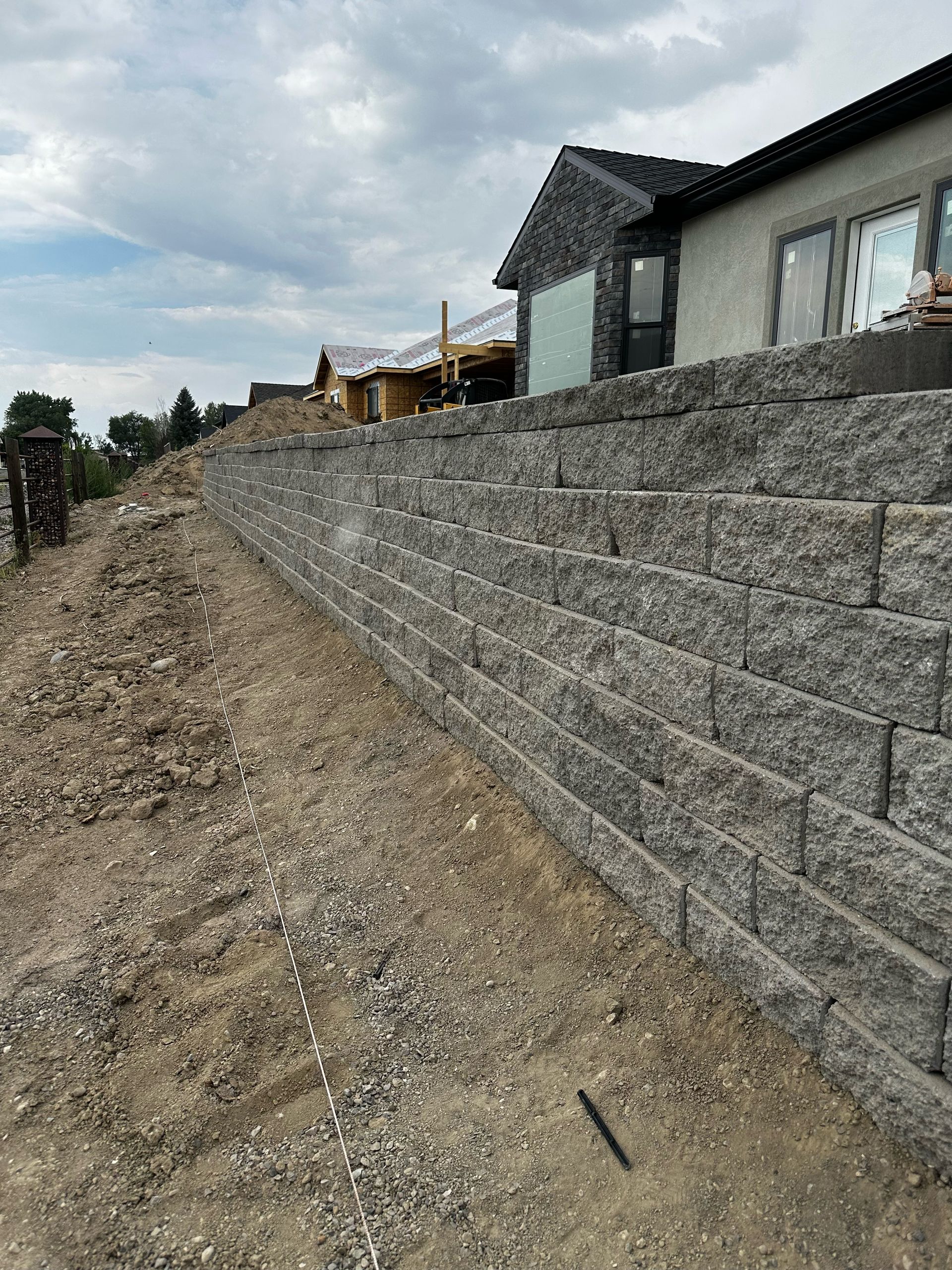 A gray retaining wall along a hillside supports a house under construction, with dirt in the foreground.