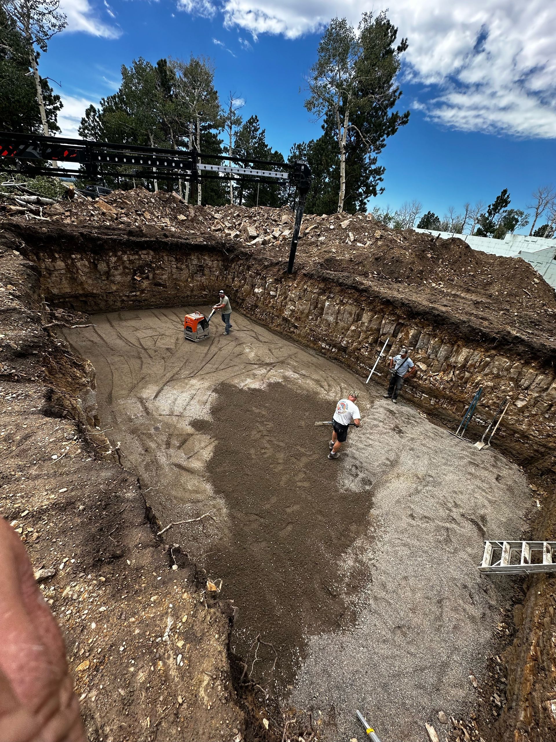 Construction site: Workers in a large excavated pit with gravel floor under blue sky with trees.