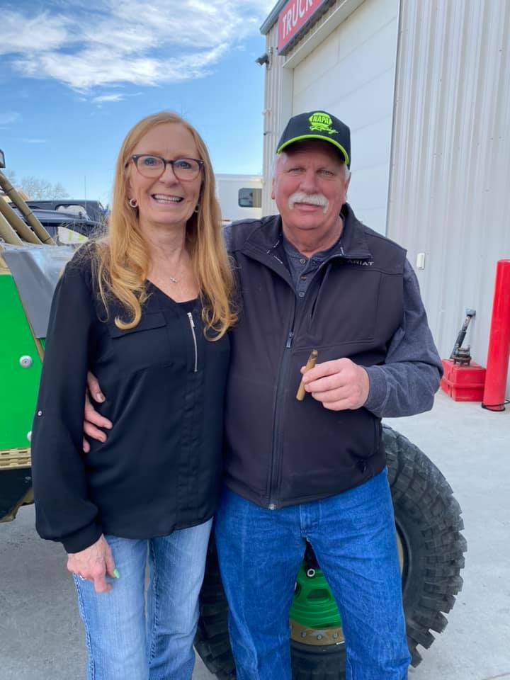 Two people standing together outdoors beside a green vehicle, smiling and posing in jackets and jeans.