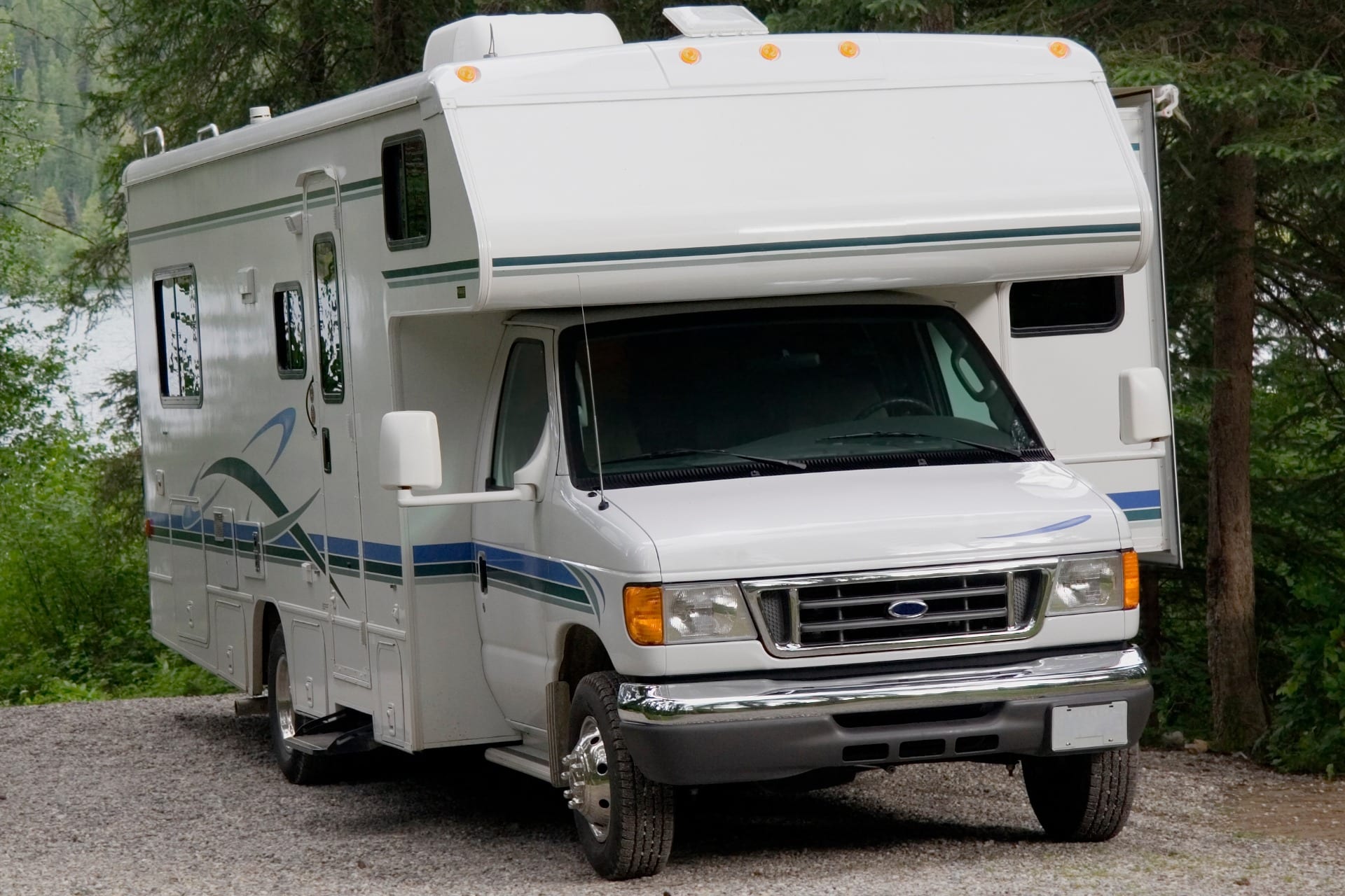White RV camper van parked on a gravel road beside green trees.