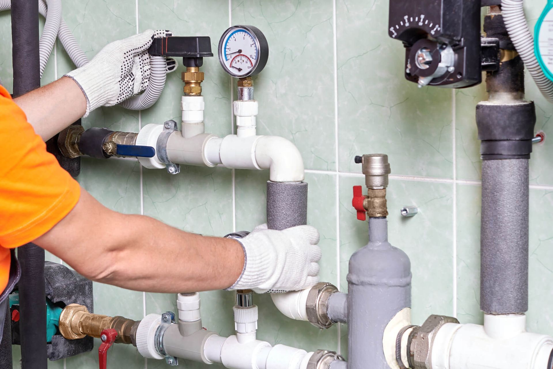 Plumber in orange shirt and gloves adjusts pipes with gauges and valves in a utility room.