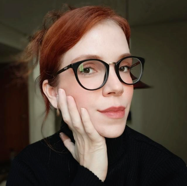 Retrato de uma pessoa ruiva de óculos pretos, com o rosto apoiado em uma das mãos contra um fundo suave.