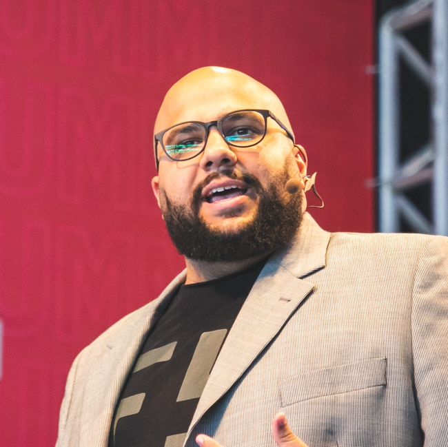 Homem discursando no palco, vestindo blazer bege e óculos, com um fundo vermelho e iluminação de treliça.