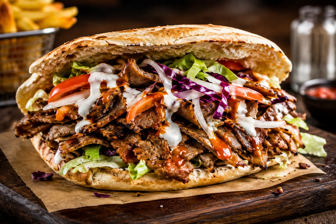 Panino pita grigliato farcito con carne, lattuga, cavolo e salsa, servito su un tagliere di legno.