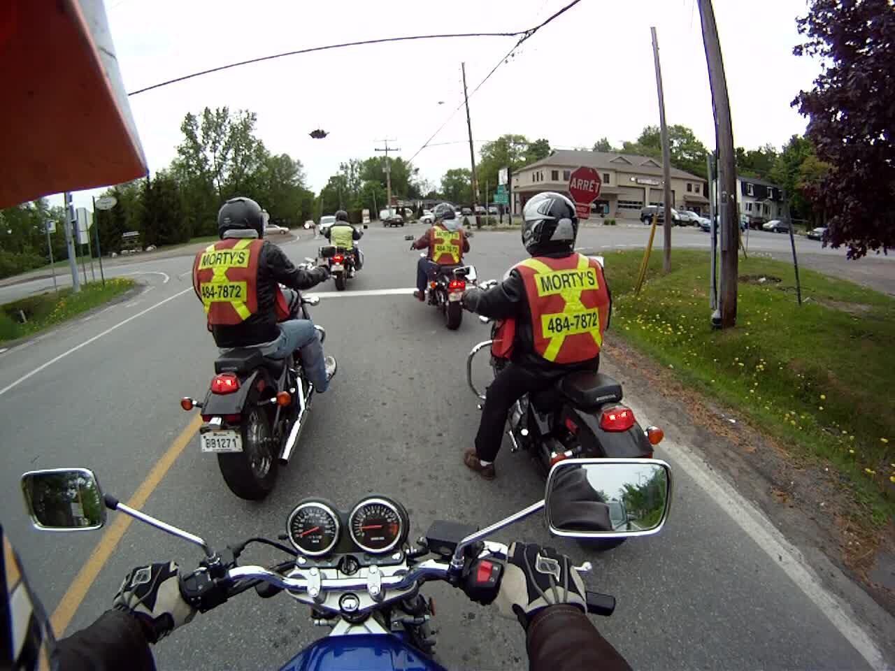 Des motards portant des gilets de sécurité se suivent sur une route avec des bâtiments en arrière-plan.