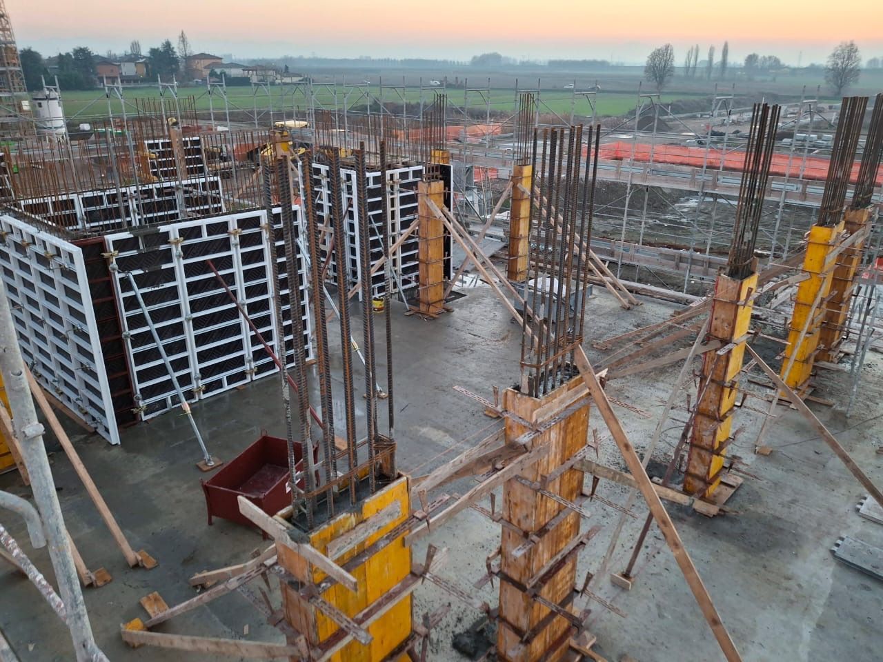 Cantiere con pilastri in cemento, casseforme gialle e impalcature all’alba.