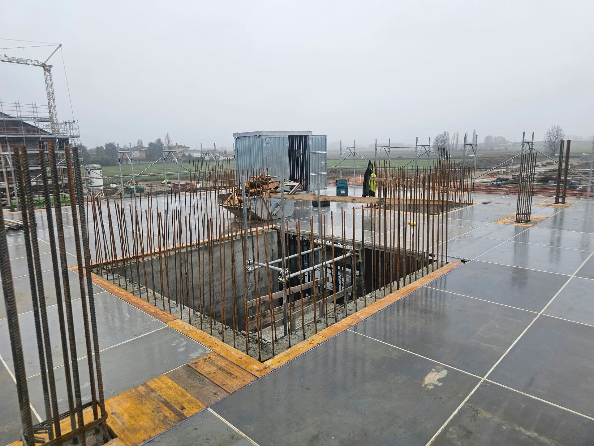 Cantiere con ferri d’armatura a vista attorno a un’apertura rettangolare nelle fondamenta, su una soletta di calcestruzzo bagnata.