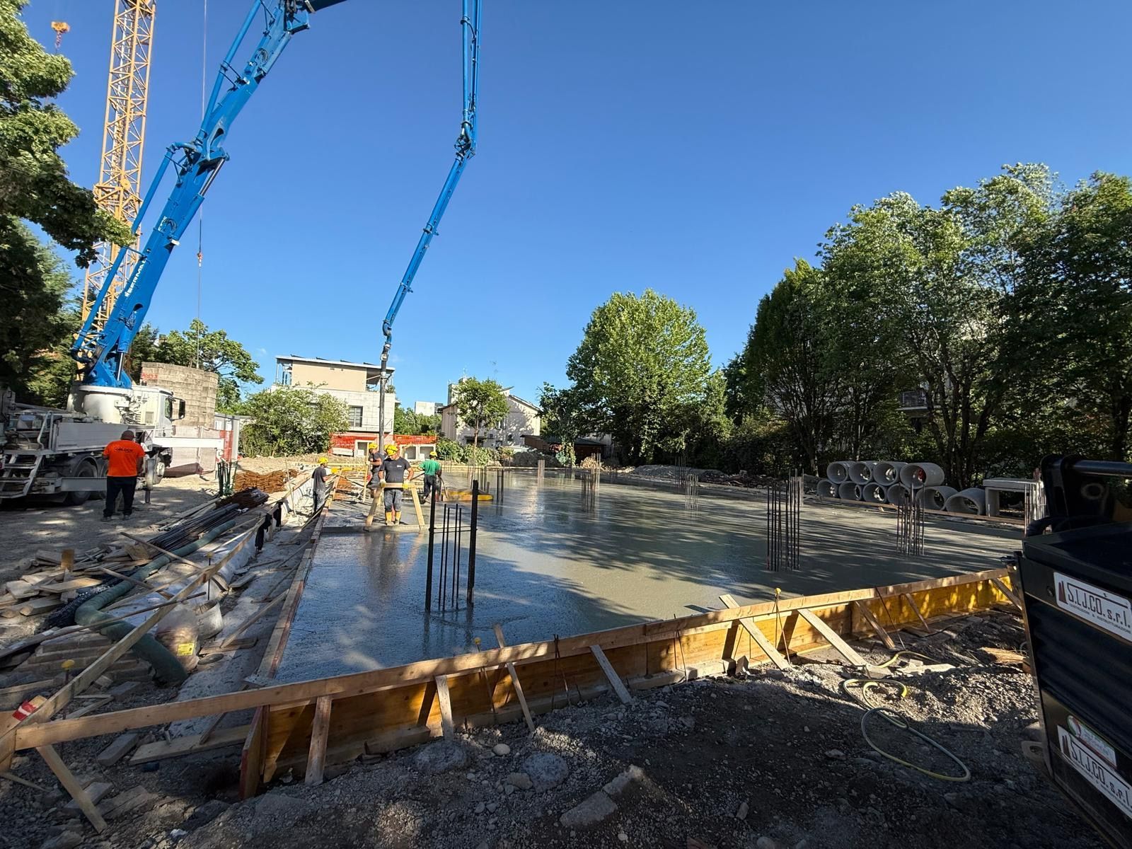 Cantiere con calcestruzzo in fase di getto, operai, bracci di pompe blu e una strada alberata sotto un cielo sereno