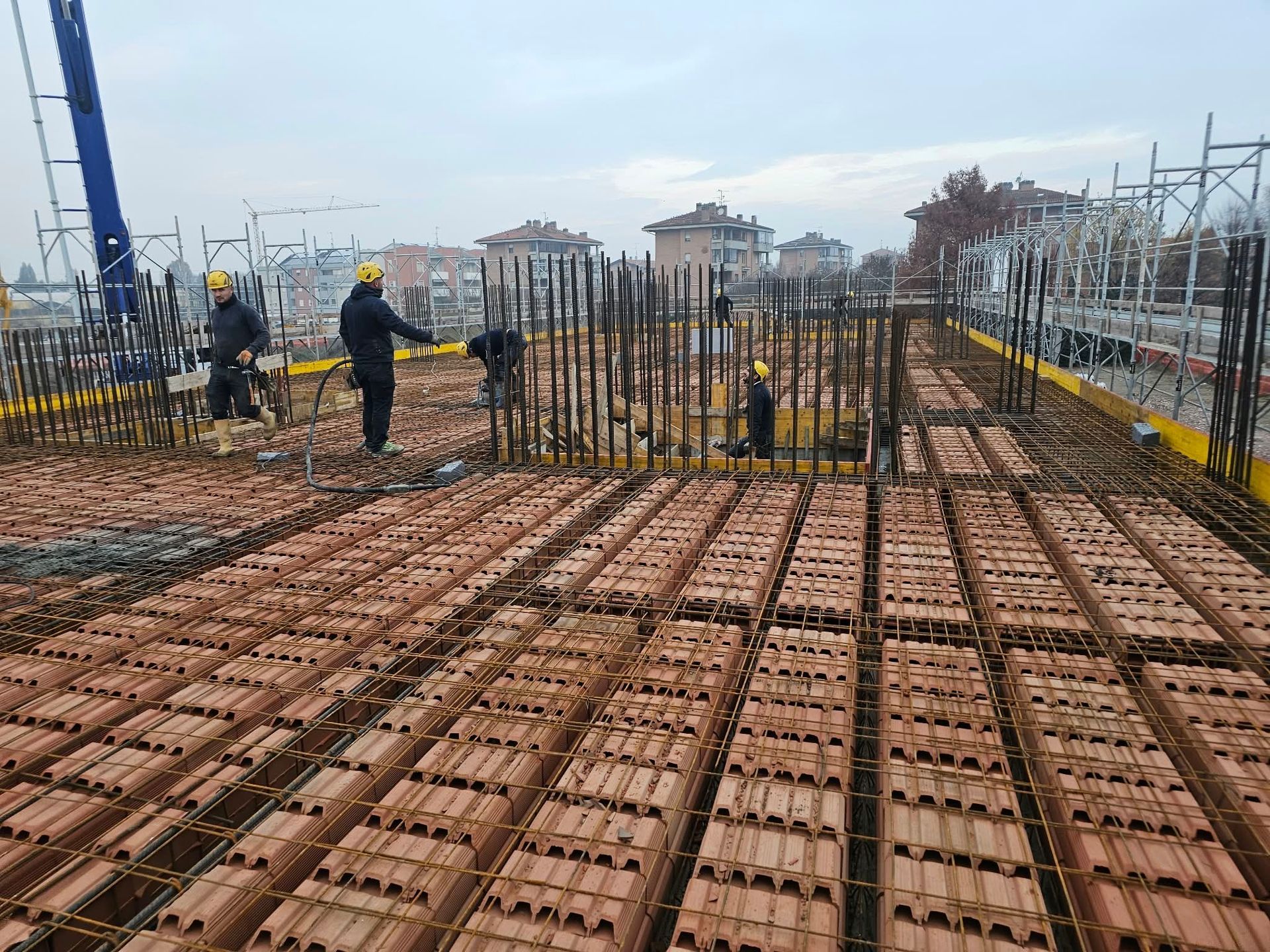 Cantiere con operai che posano le barre d’armatura su un pavimento di blocchi rossi, sotto un cielo coperto
