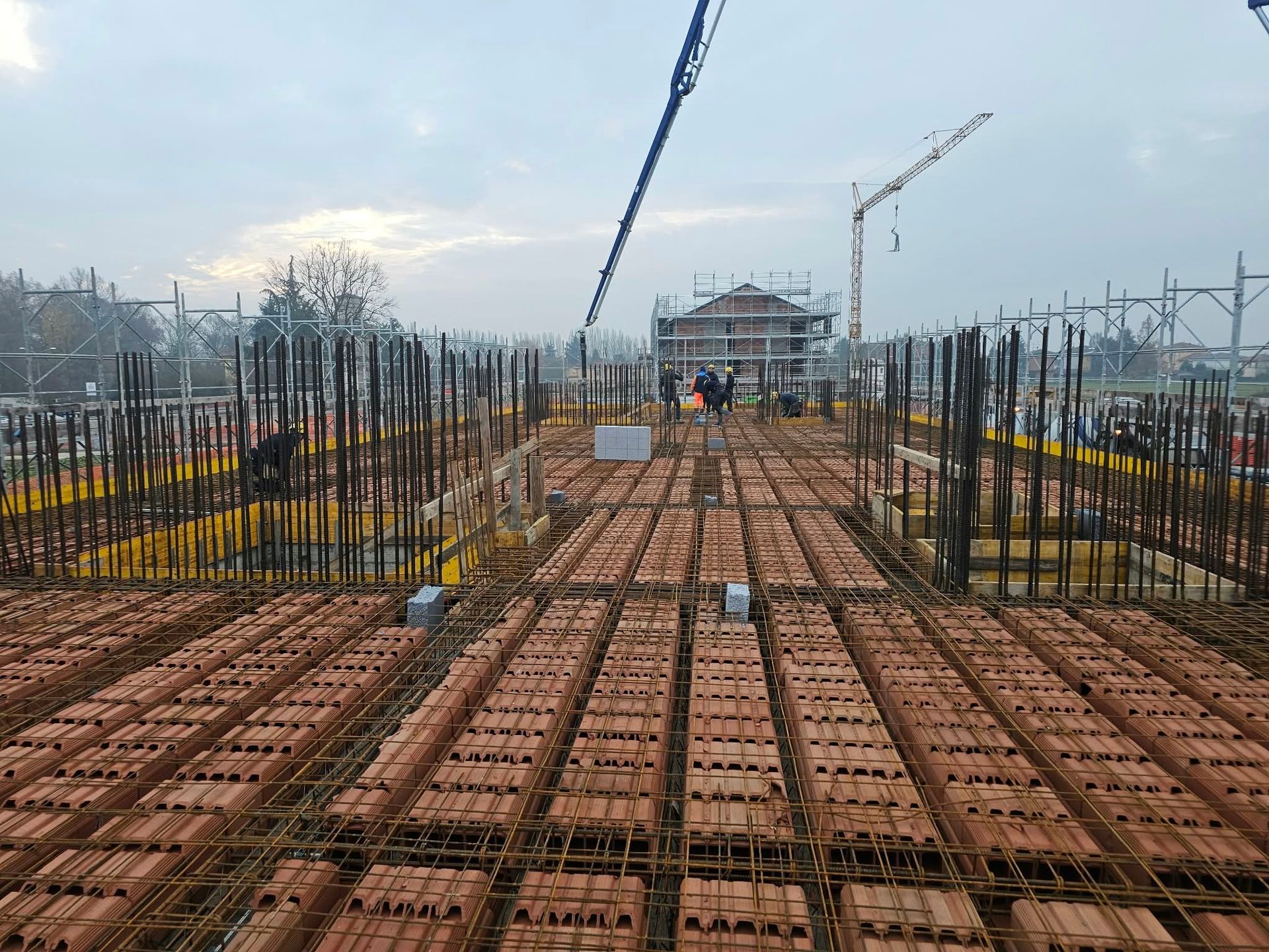 Cantiere con fondamenta a griglia di tondini d’armatura, casseforme in calcestruzzo e una gru sotto un cielo nuvoloso