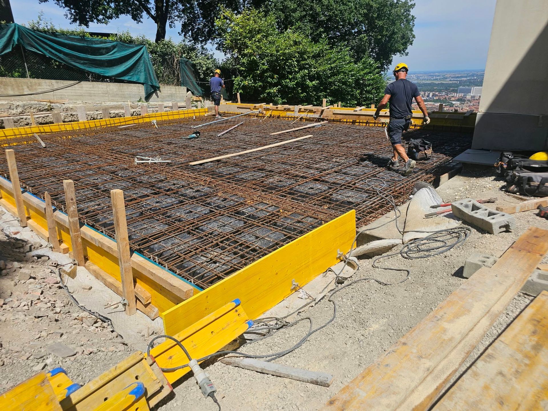 Operai al lavoro su una fondazione in cemento armato in un cantiere in collina, con casseforme gialle e cemento nelle vicinanze