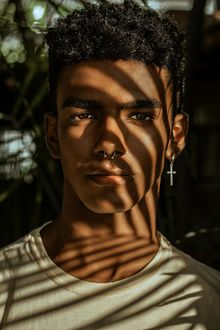 Portrait of a person in a white shirt, lit by striped window shadows, with a dangling cross earring