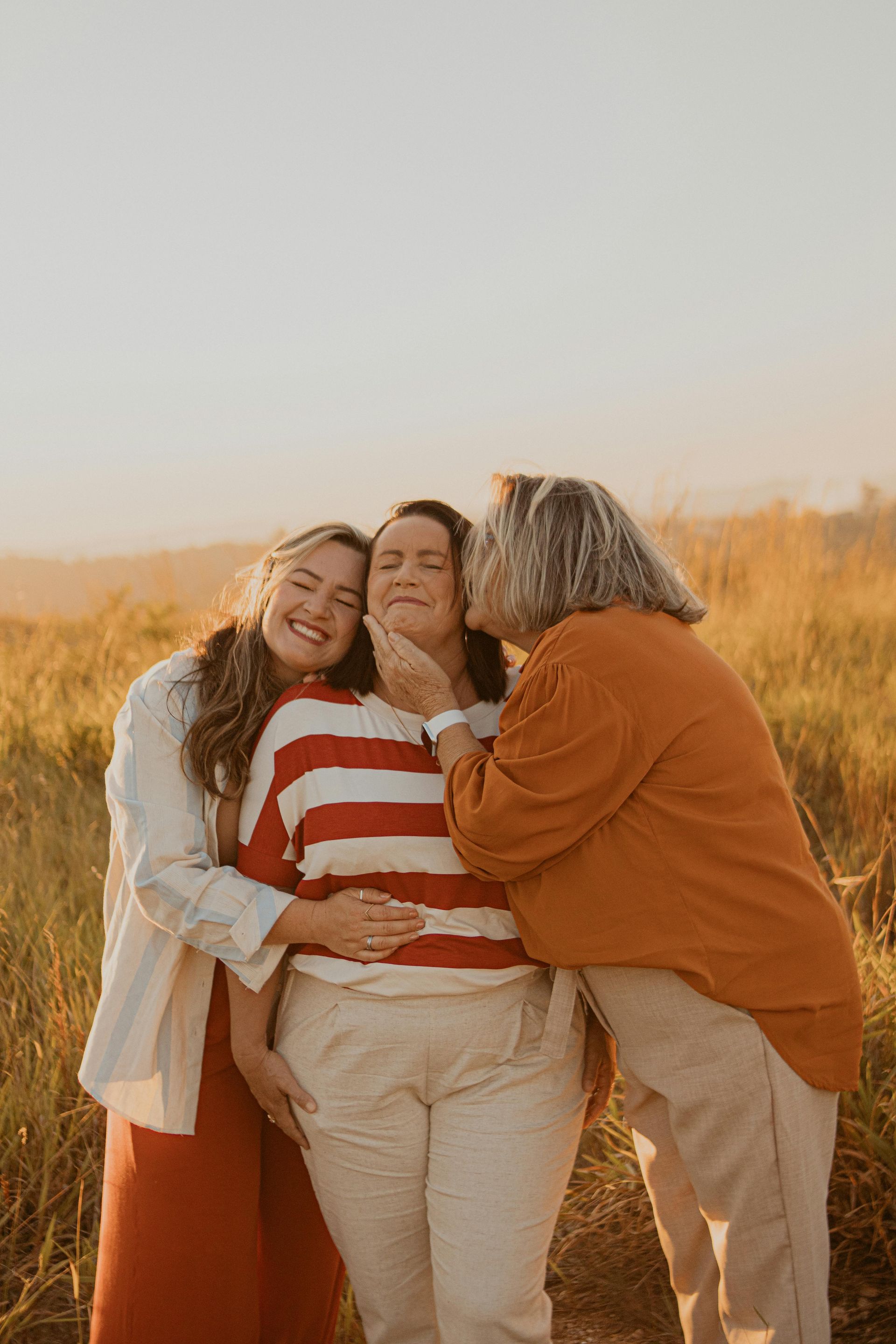 Three people hugging in a sunlit field, smiling and leaning together.
