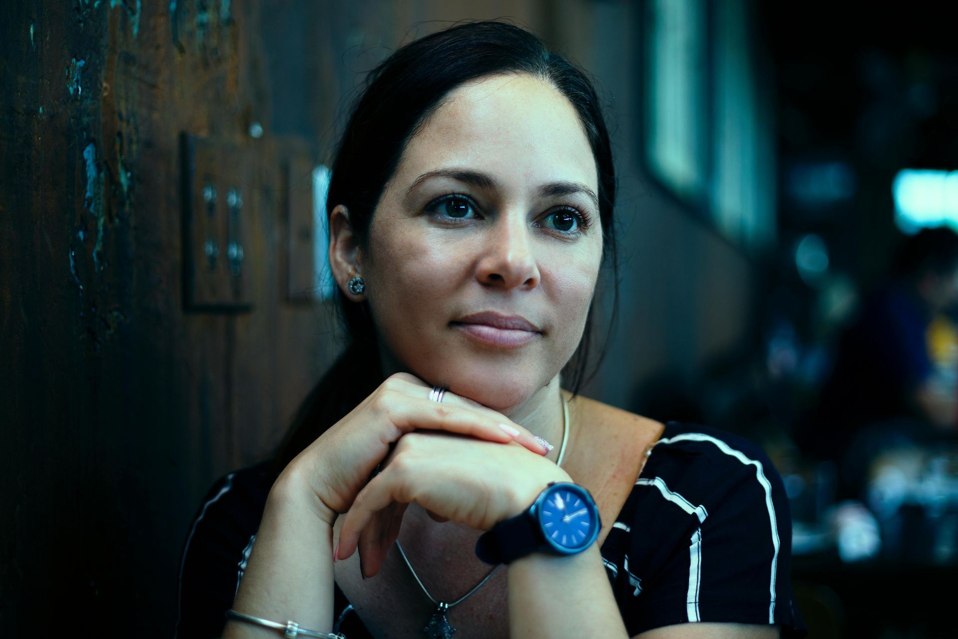 Woman resting chin on clasped hands, wearing a blue watch in a dim café setting.