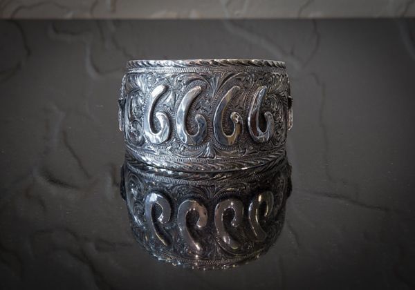 Silver cuff bracelet with ornate, swirling patterns, reflected on a dark, textured surface.