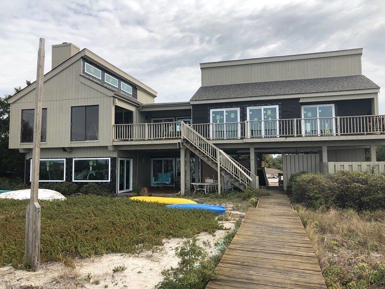 Beach house with wooden boardwalk, deck, and kayaks on the sand. Cloudy sky overhead.