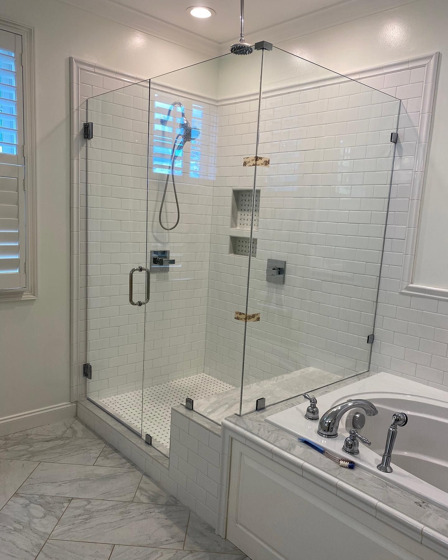 Glass shower enclosure in a white-tiled bathroom with a bathtub.