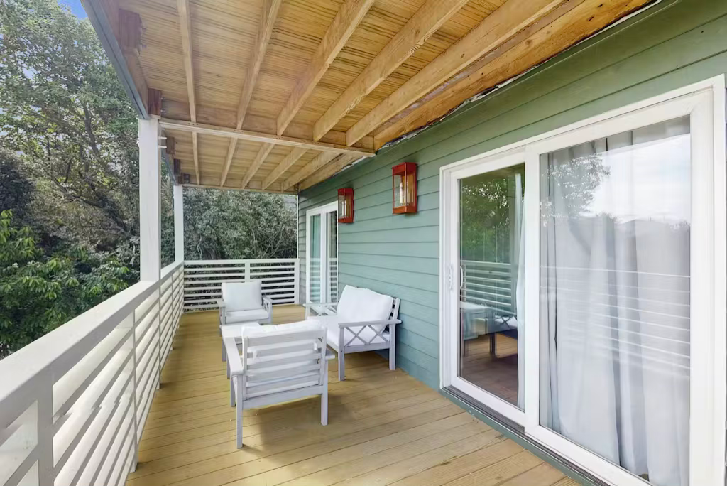 Balcony with white furniture, wood flooring, and green siding. Trees in the background, sliding glass door.