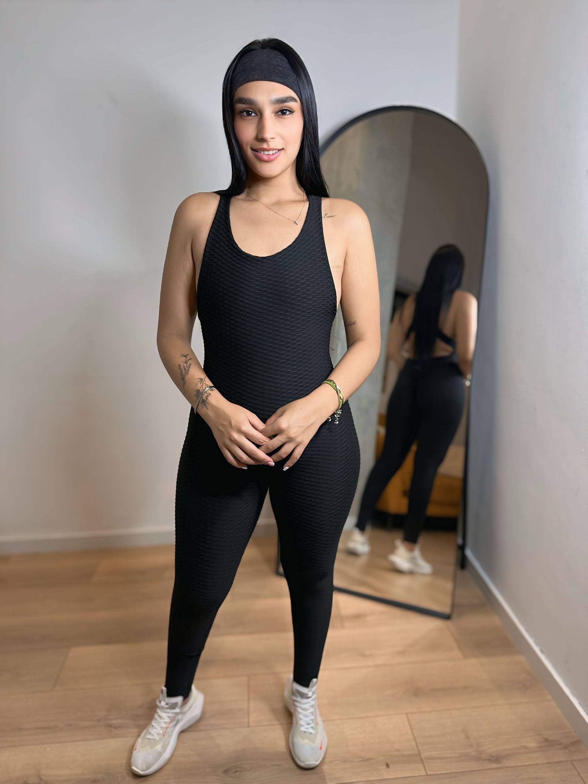 A woman in a black jumpsuit is standing in front of a mirror.