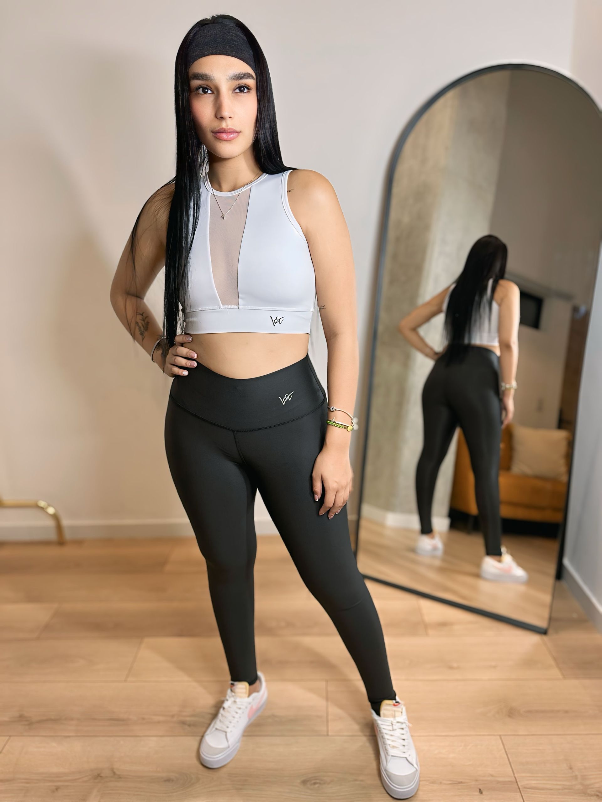A woman in a white crop top and black leggings is standing in front of a mirror.