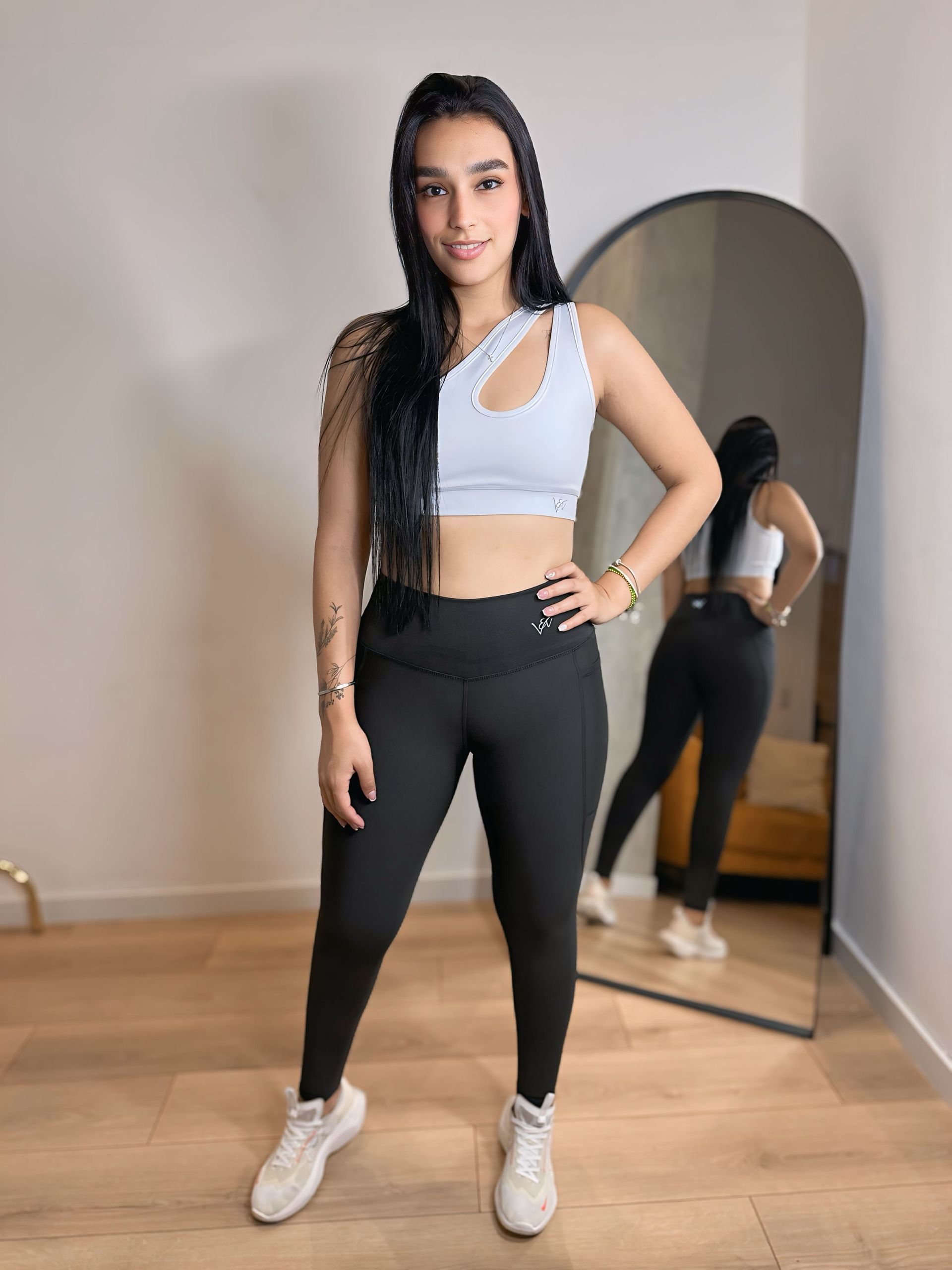 A woman in a white crop top and black leggings is standing in front of a mirror.