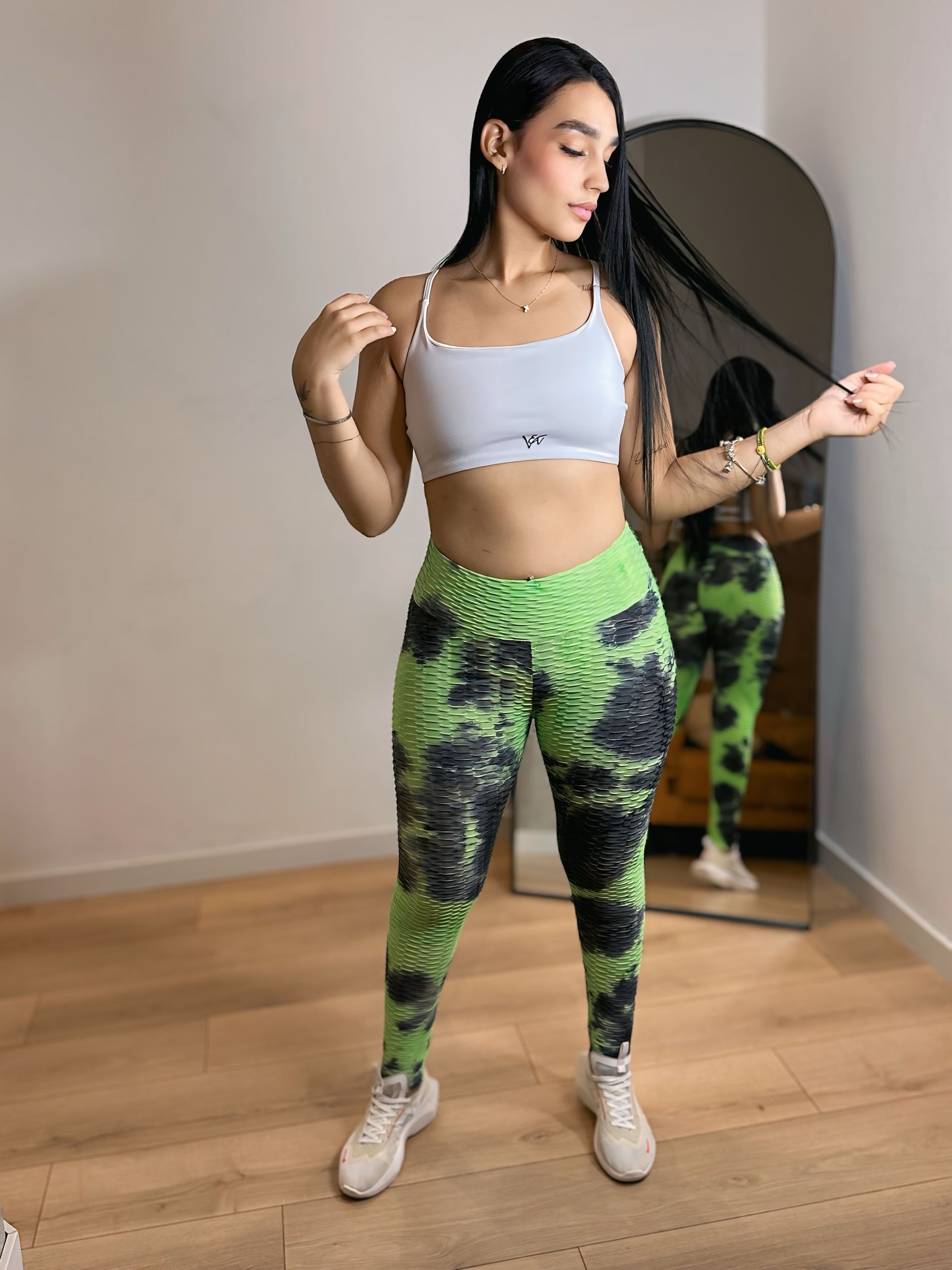 A woman in a white top and green leggings is standing in front of a mirror.