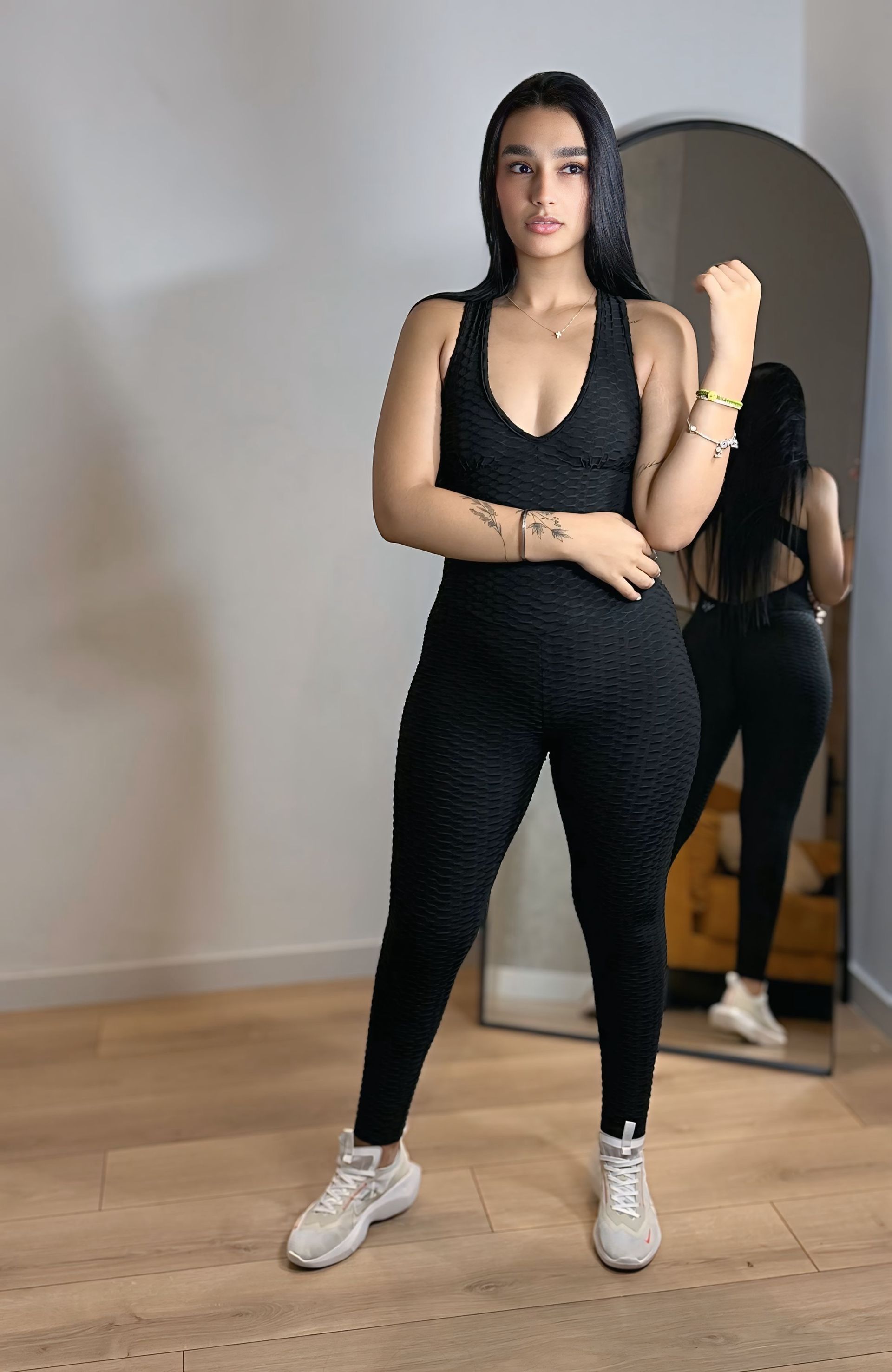 A woman in a black jumpsuit is standing in front of a mirror.