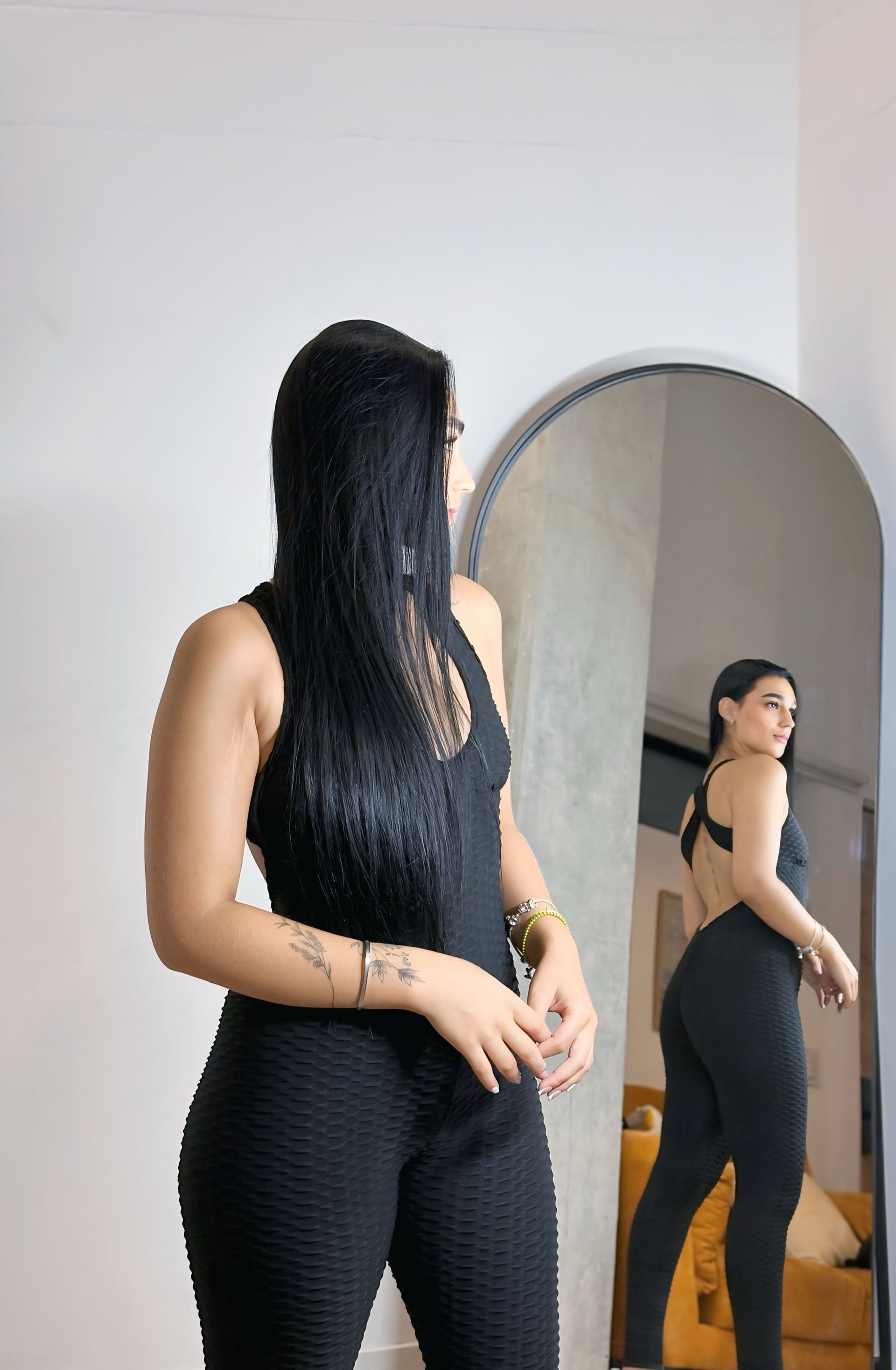 A woman in a black jumpsuit is standing in front of a mirror.
