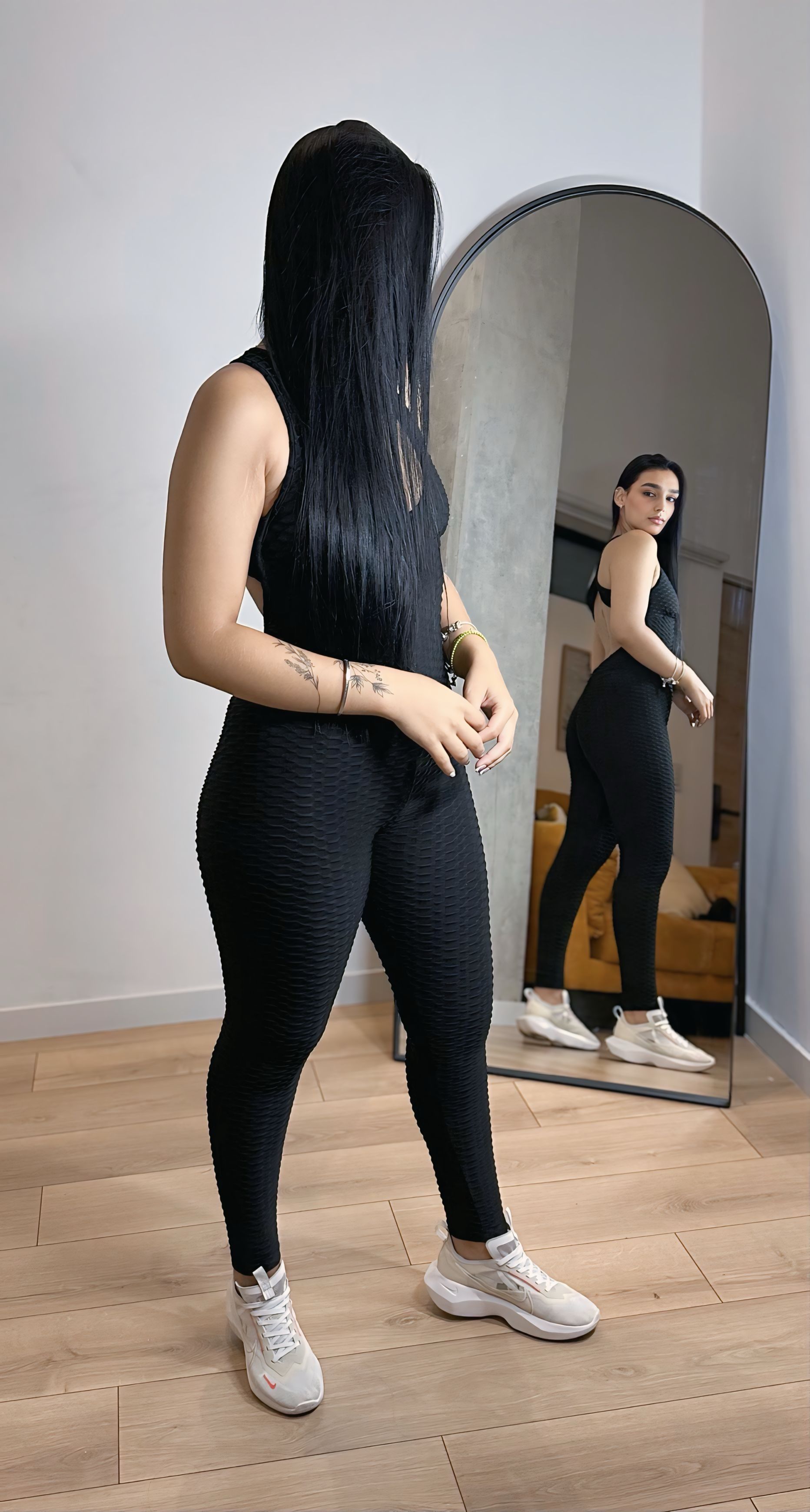 A woman in a black jumpsuit is standing in front of a mirror.