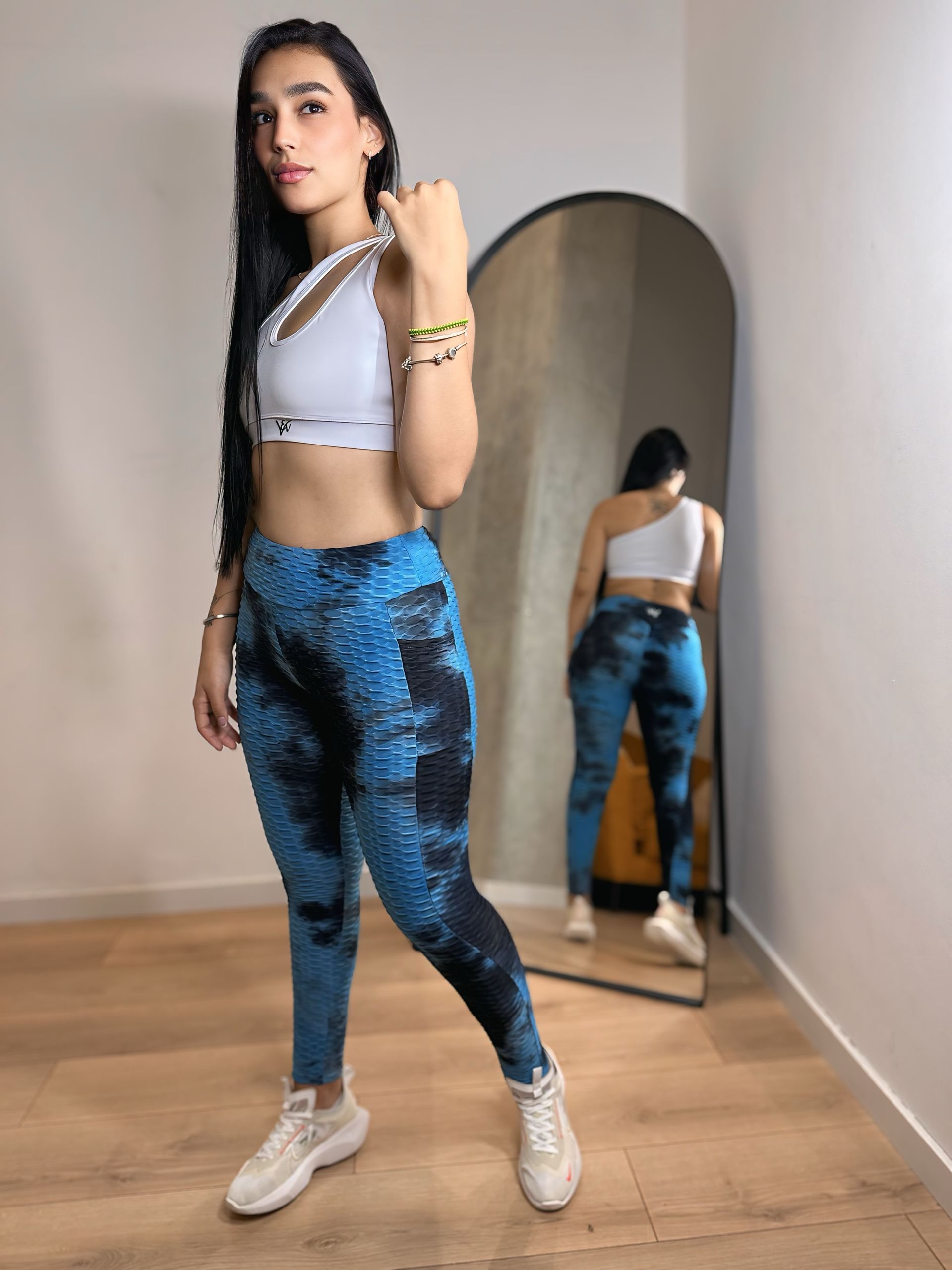 A woman in a white top and blue leggings is standing in front of a mirror.