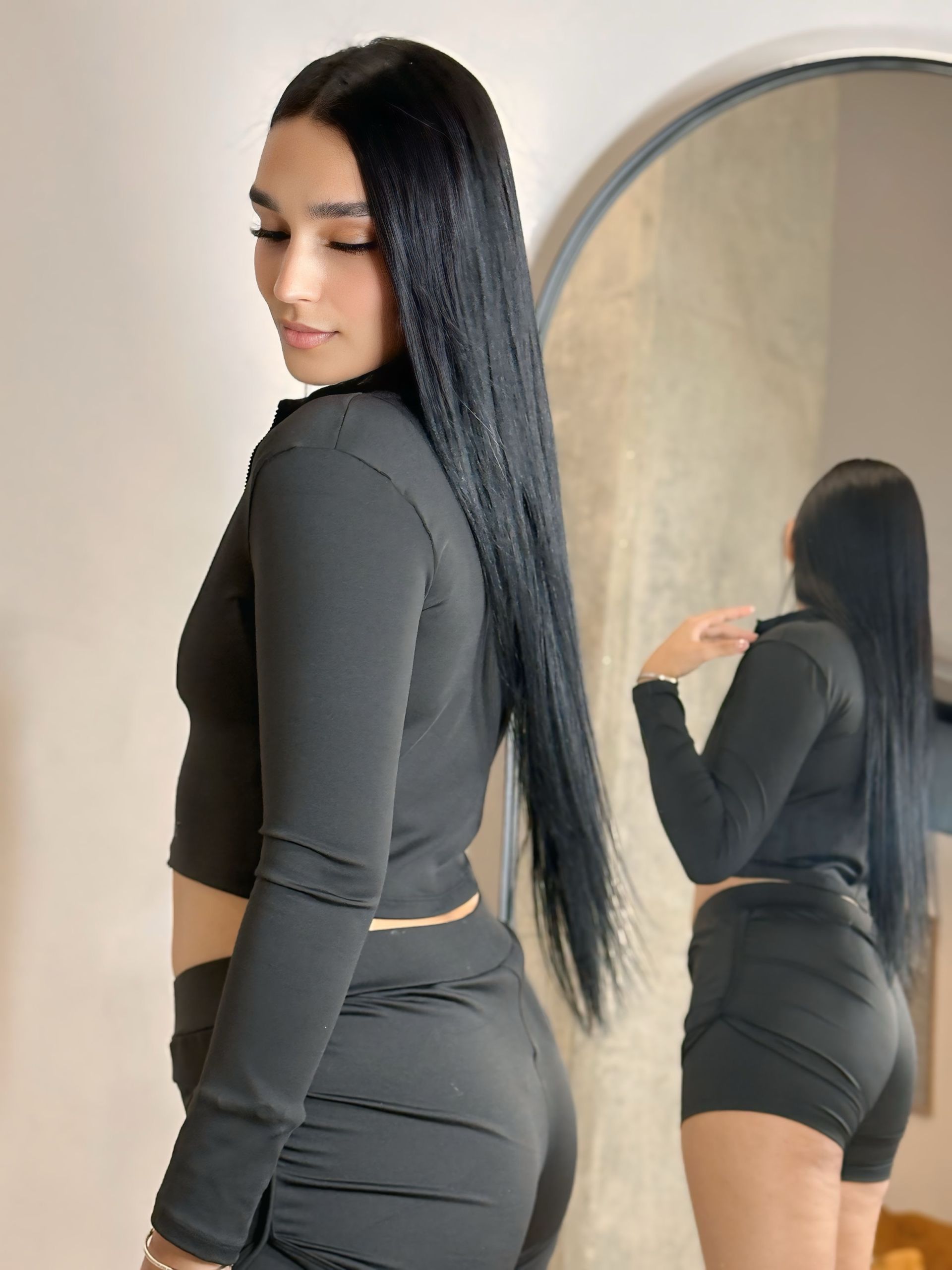 A woman in a black top and shorts is standing in front of a mirror.