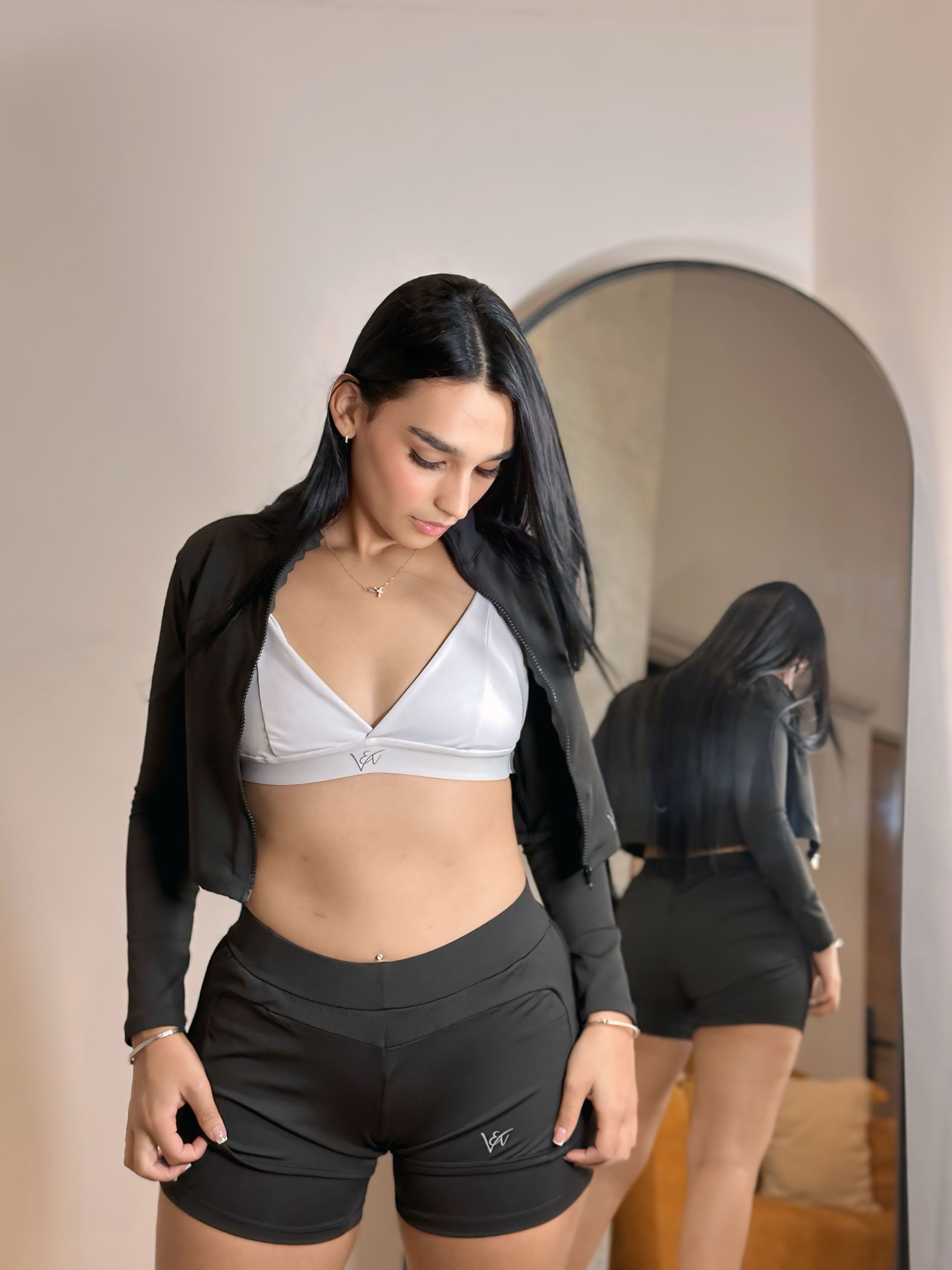 A woman in a white bra and black shorts is standing in front of a mirror.