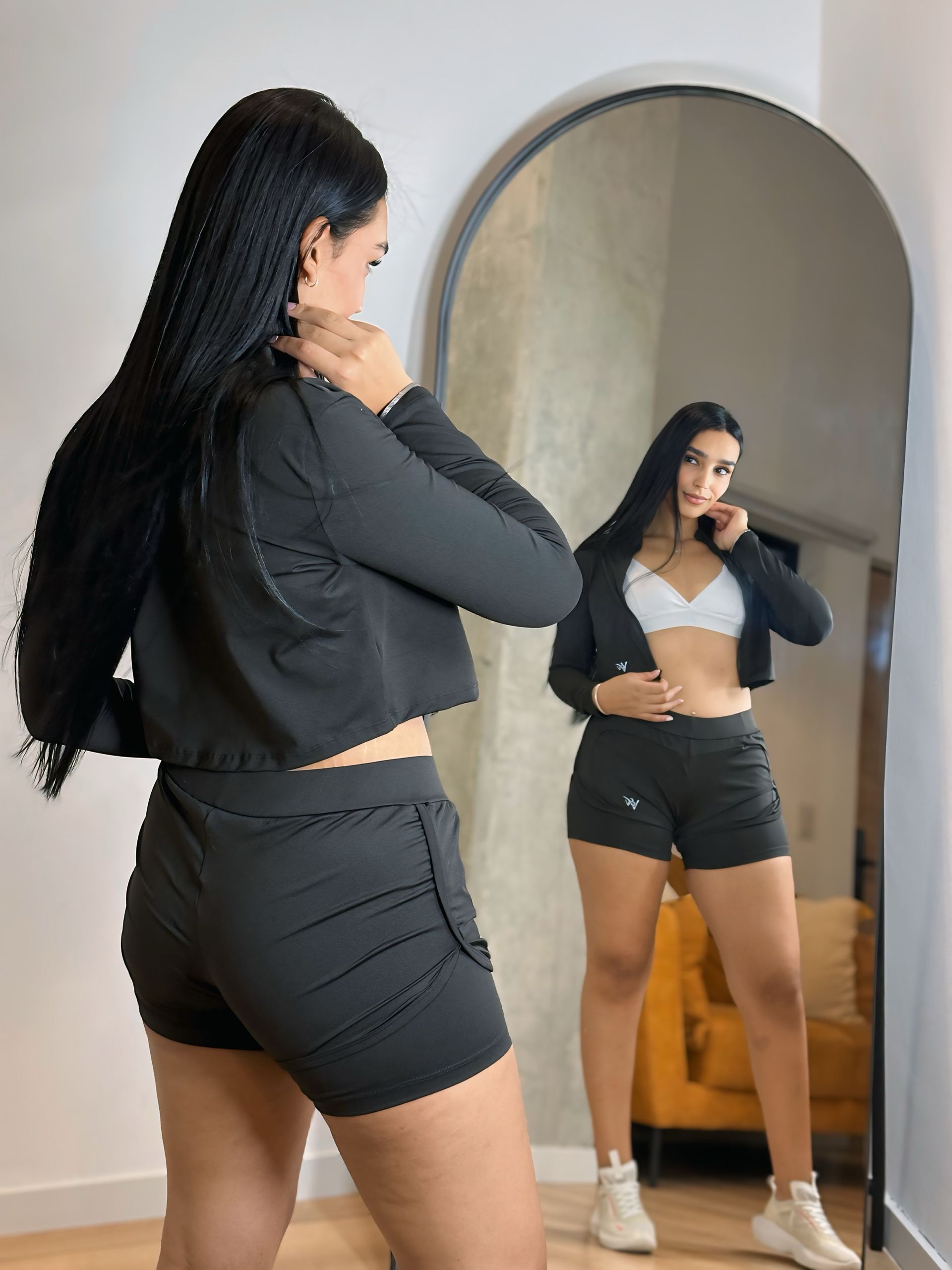 A woman is standing in front of a mirror wearing shorts and a crop top.
