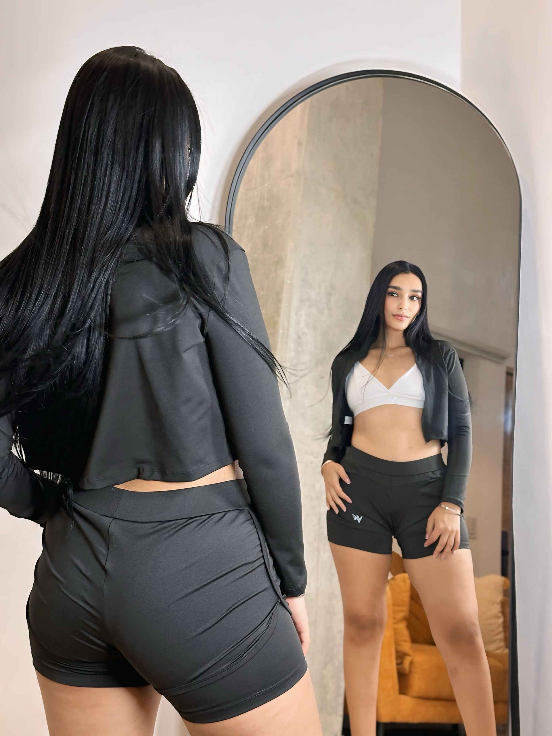 A woman in a crop top and shorts is standing in front of a mirror.
