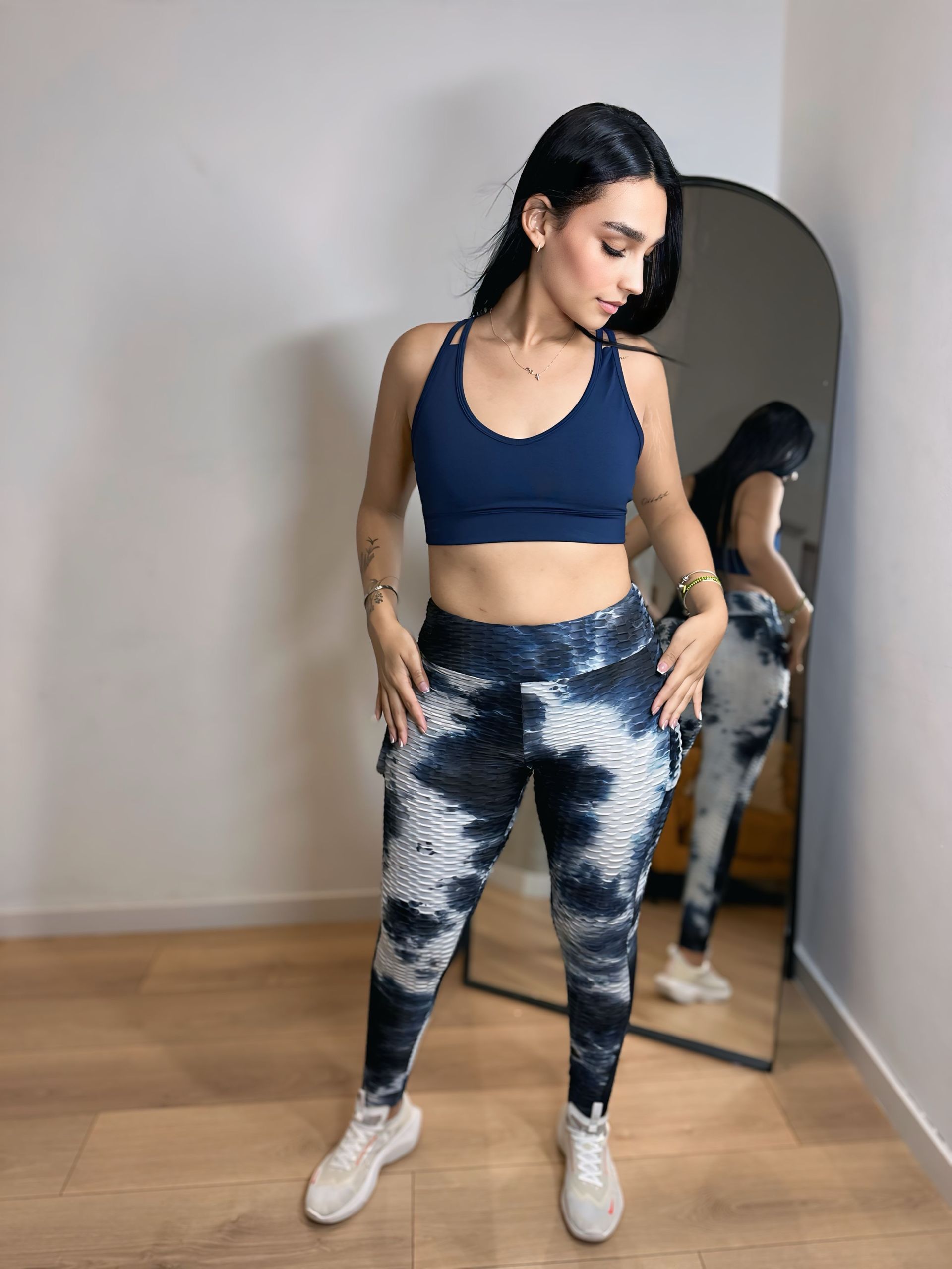 A woman in a blue sports bra and tie dye leggings is standing in front of a mirror.