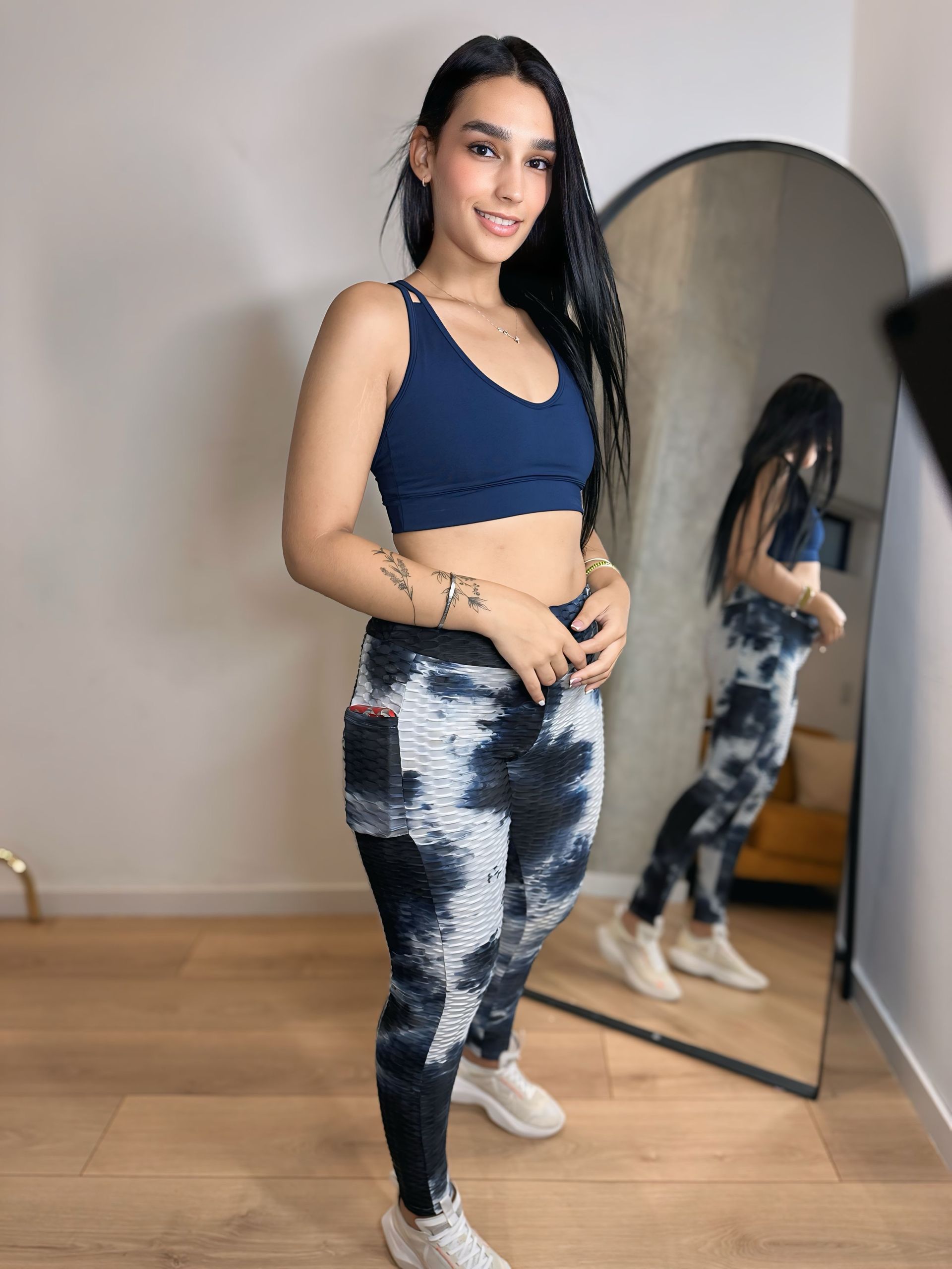 A woman in a blue crop top and tie dye leggings is standing in front of a mirror.