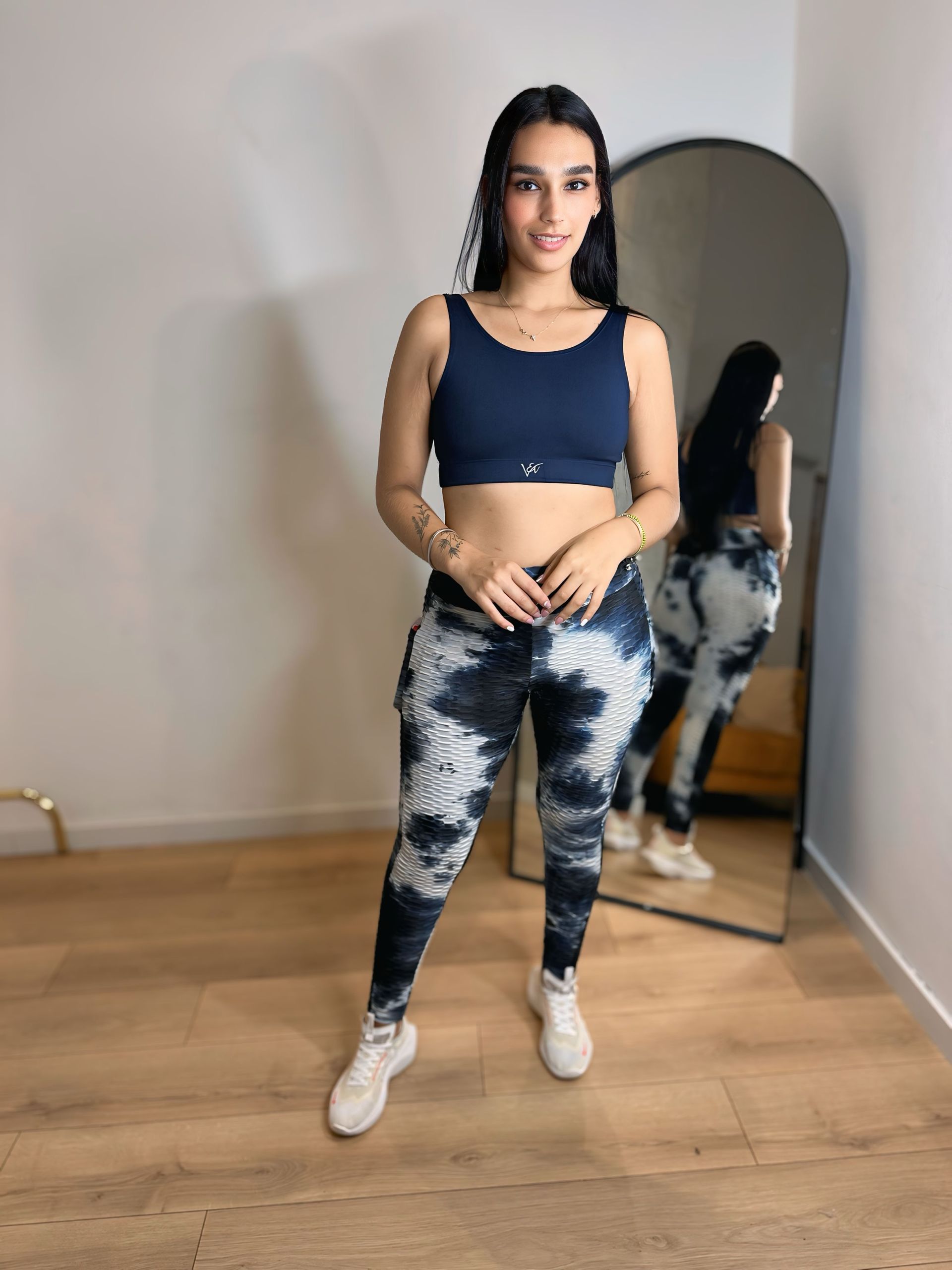 A woman in a crop top and tie dye leggings is standing in front of a mirror.