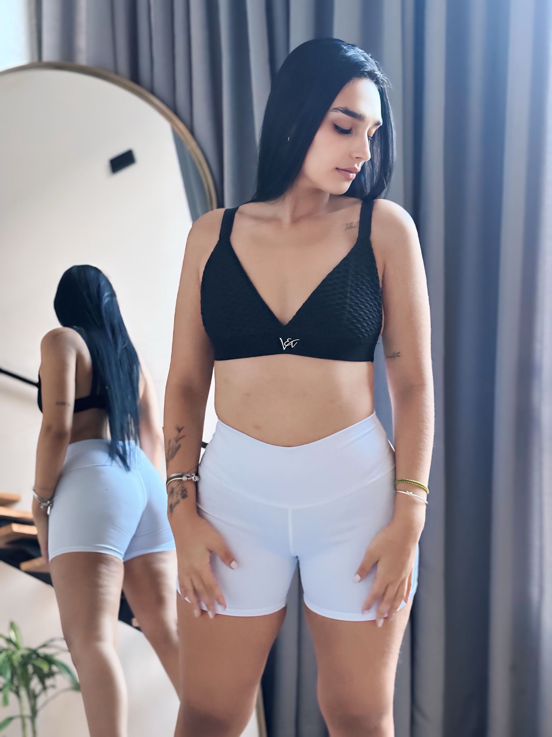 A woman in a black bra and white shorts is standing in front of a mirror.