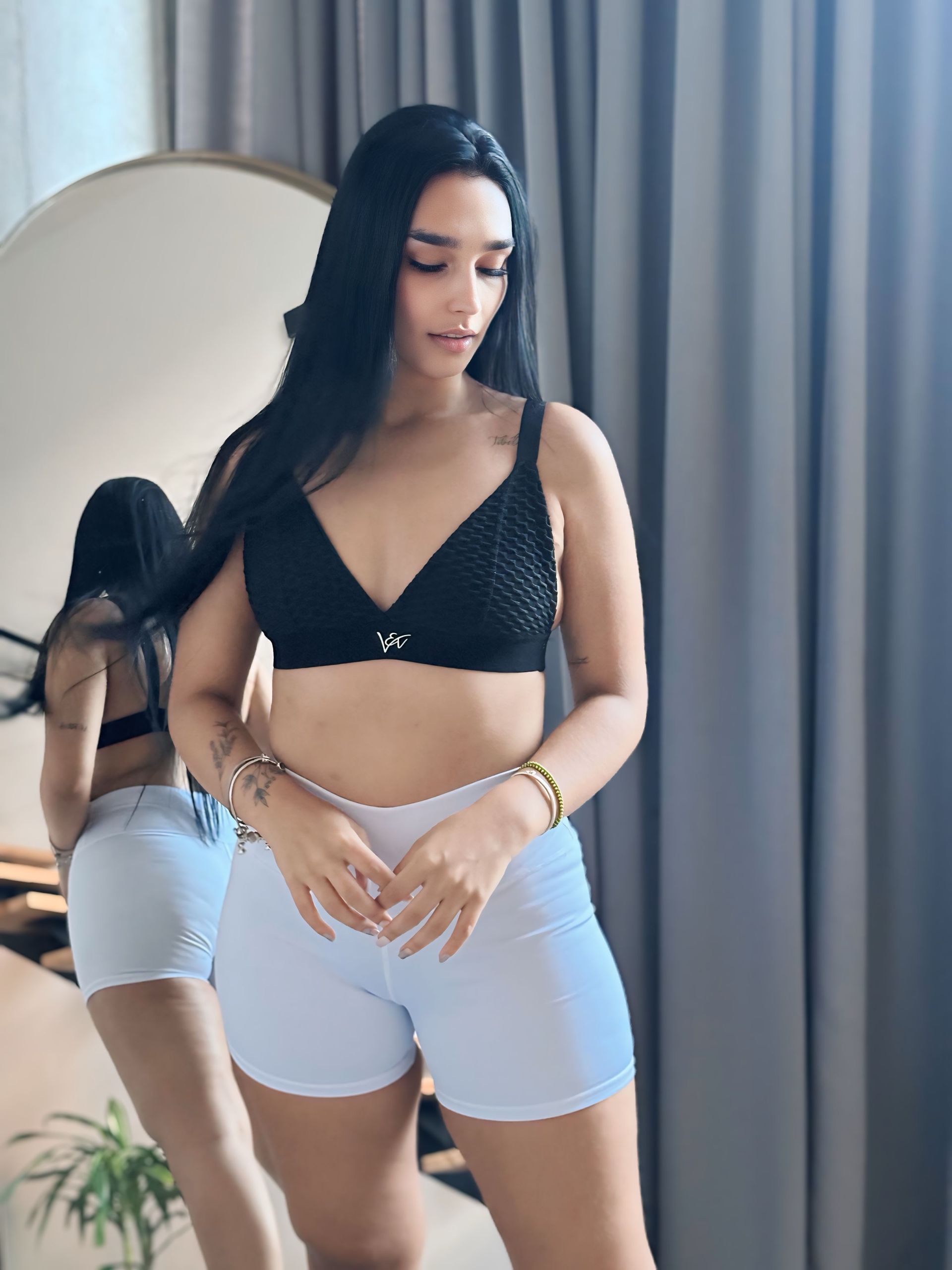 A woman in a black bra and white shorts is standing in front of a mirror.