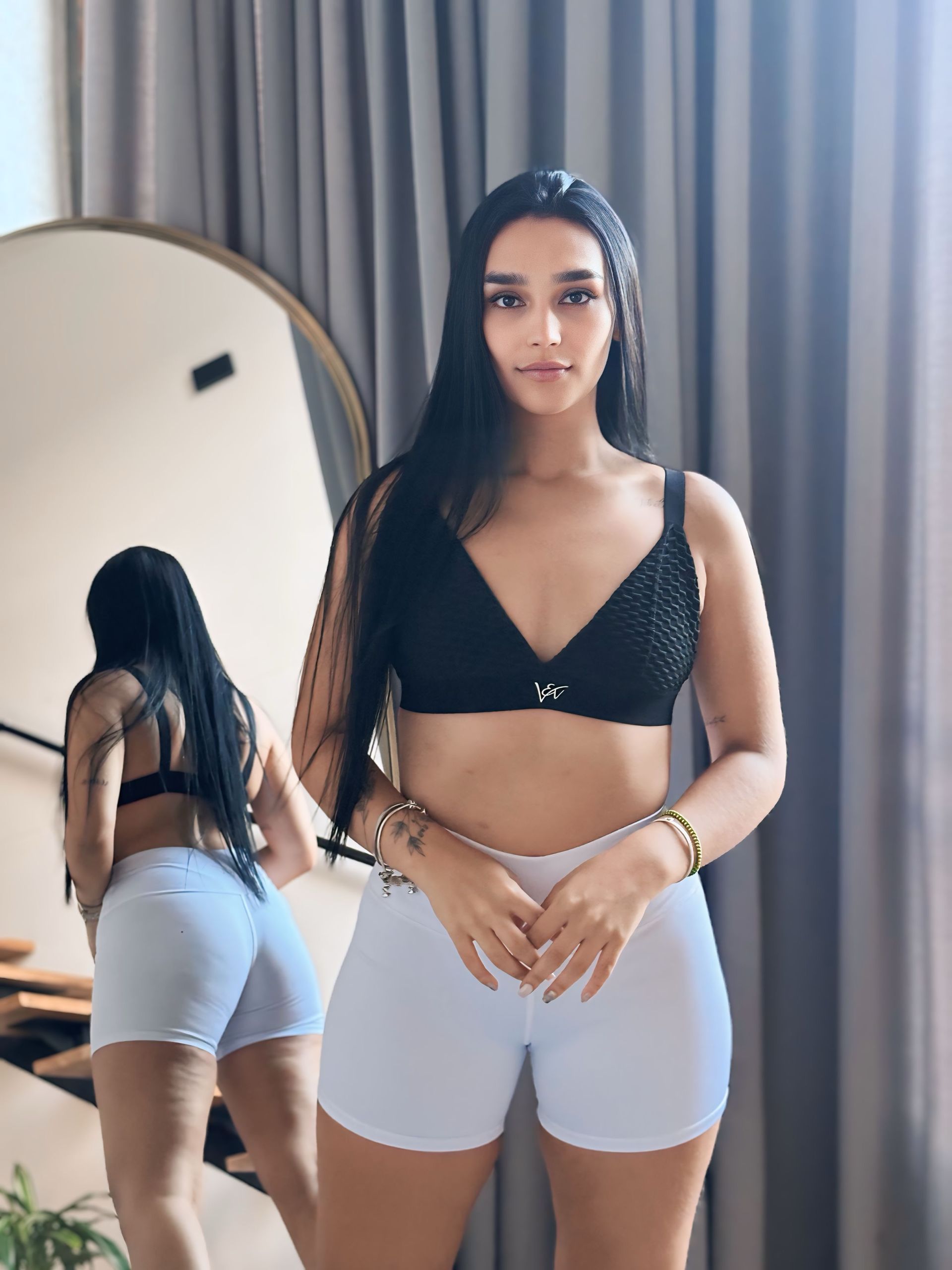 A woman in a black bra and white shorts is standing in front of a mirror.