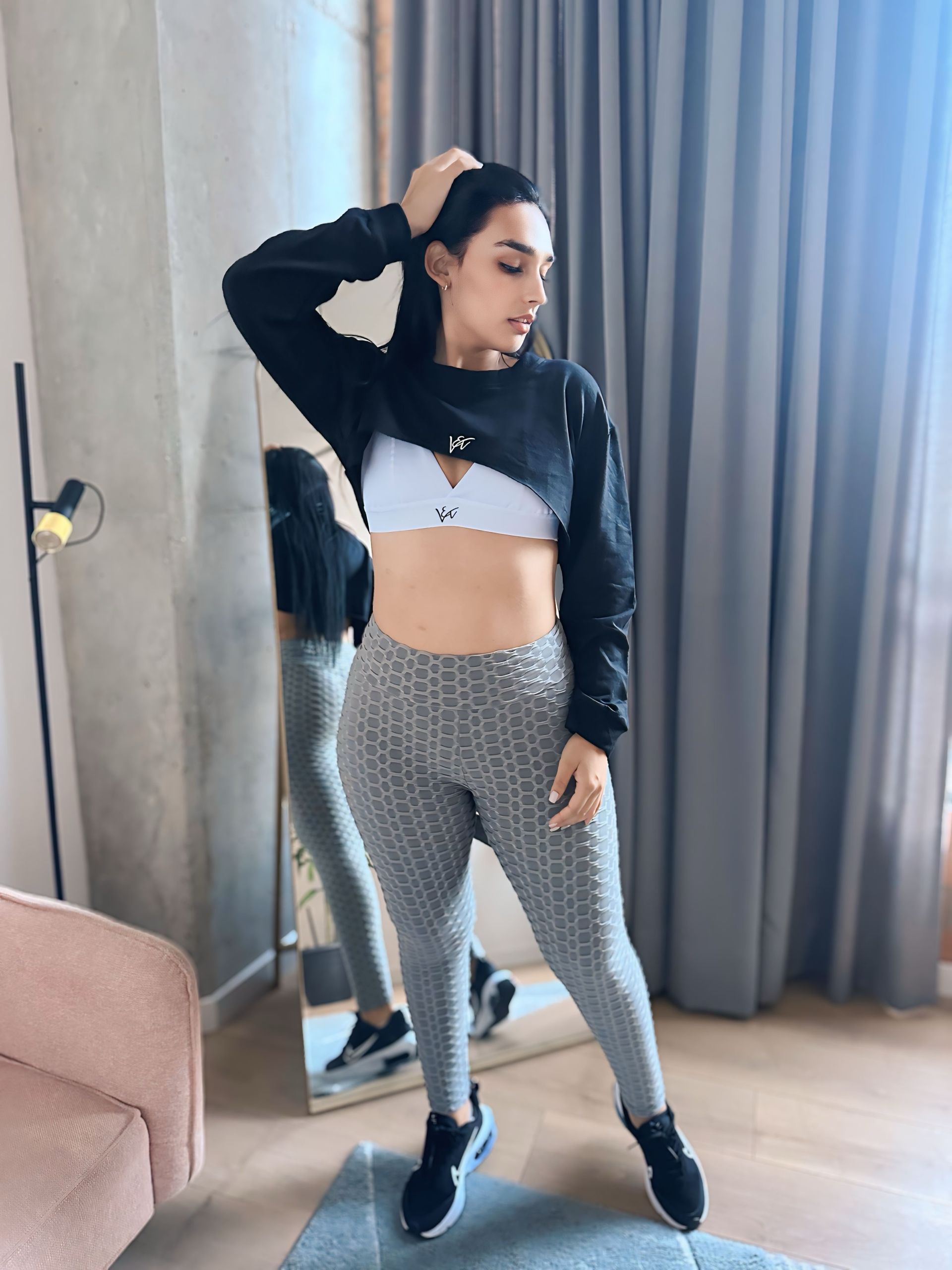 A woman in a crop top and leggings is standing in front of a mirror.