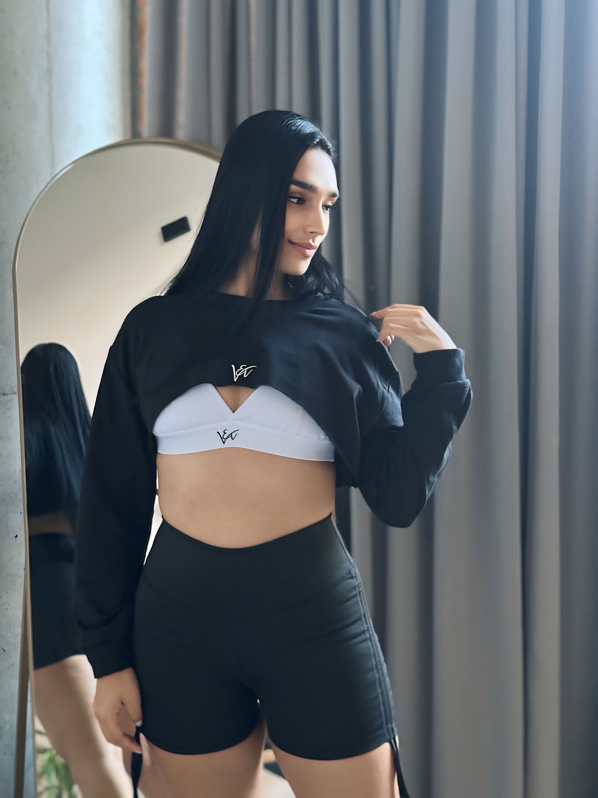 A woman in a black crop top and black shorts is standing in front of a mirror.