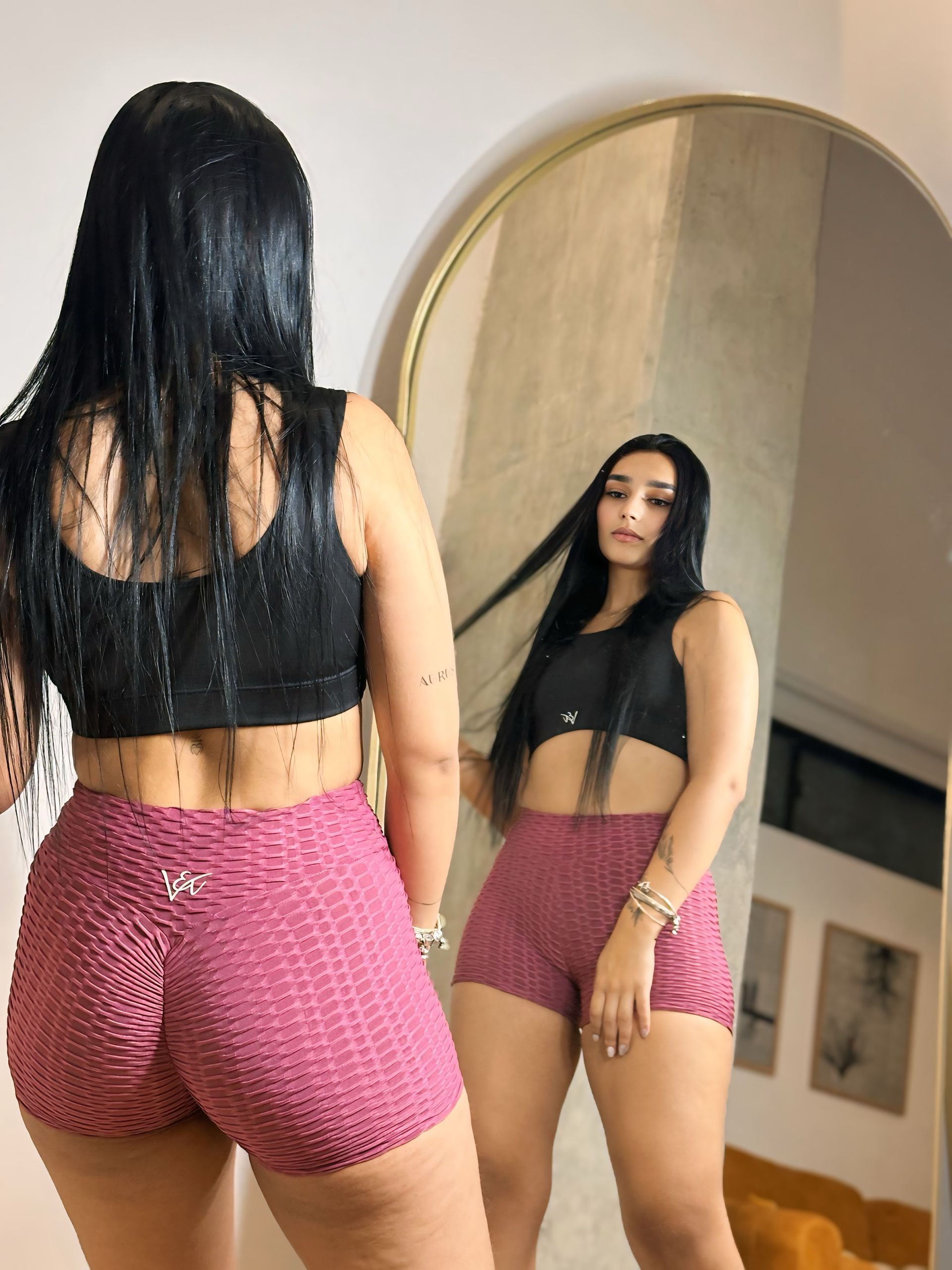 A woman in a black top and pink shorts is standing in front of a mirror.
