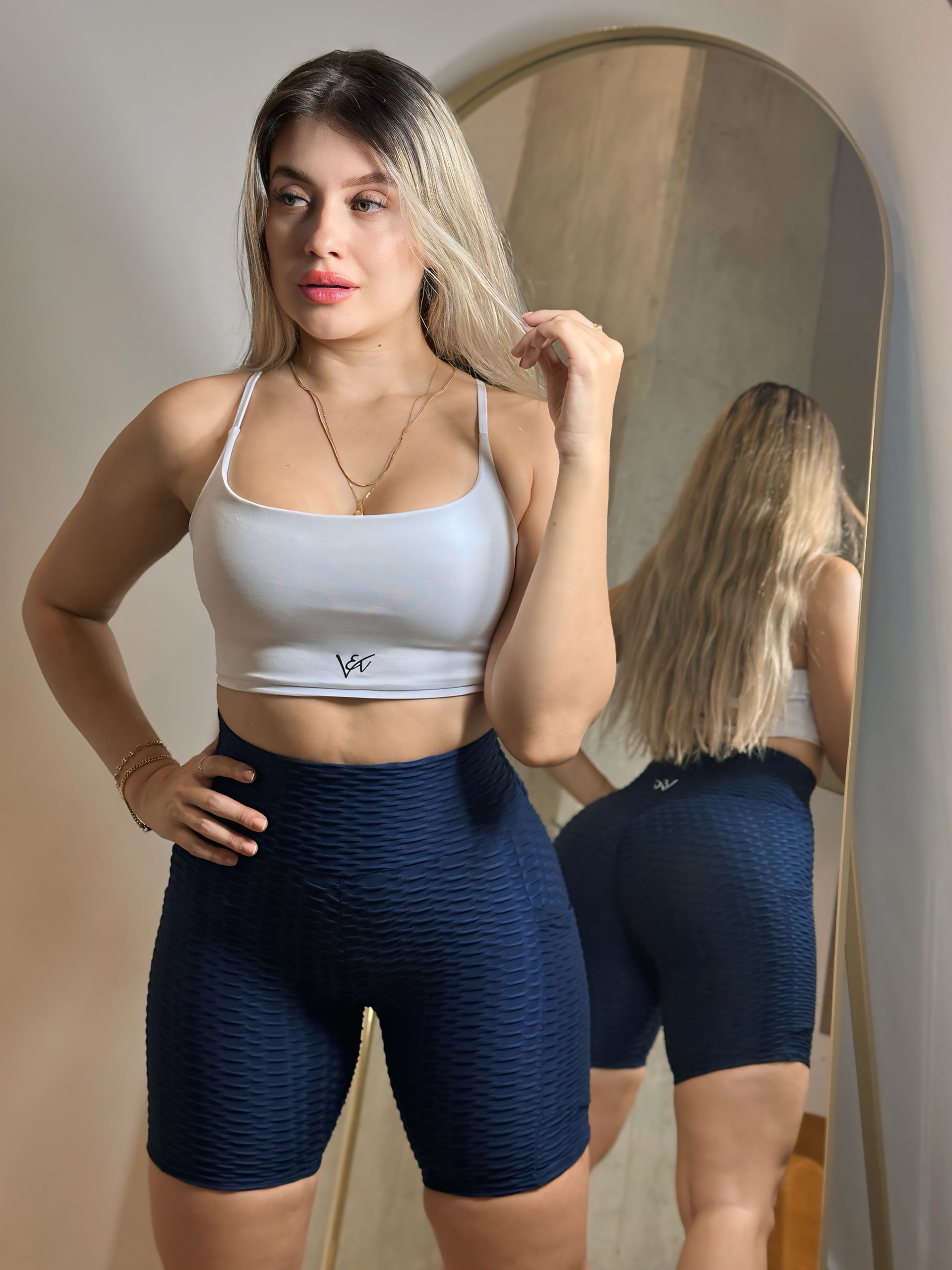 A woman in a white top and blue shorts is standing in front of a mirror.