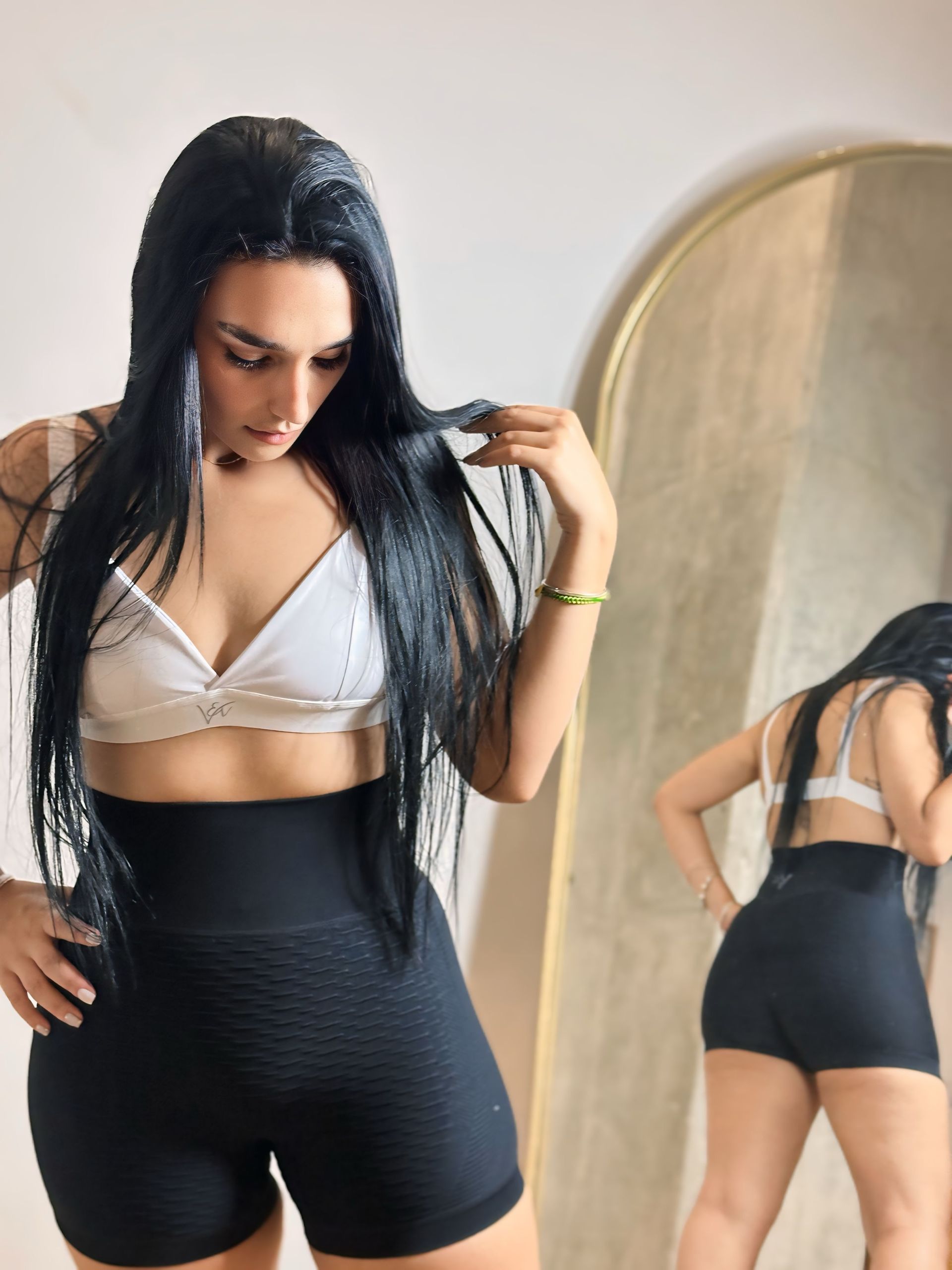 A woman in a white bra and black shorts is standing in front of a mirror.